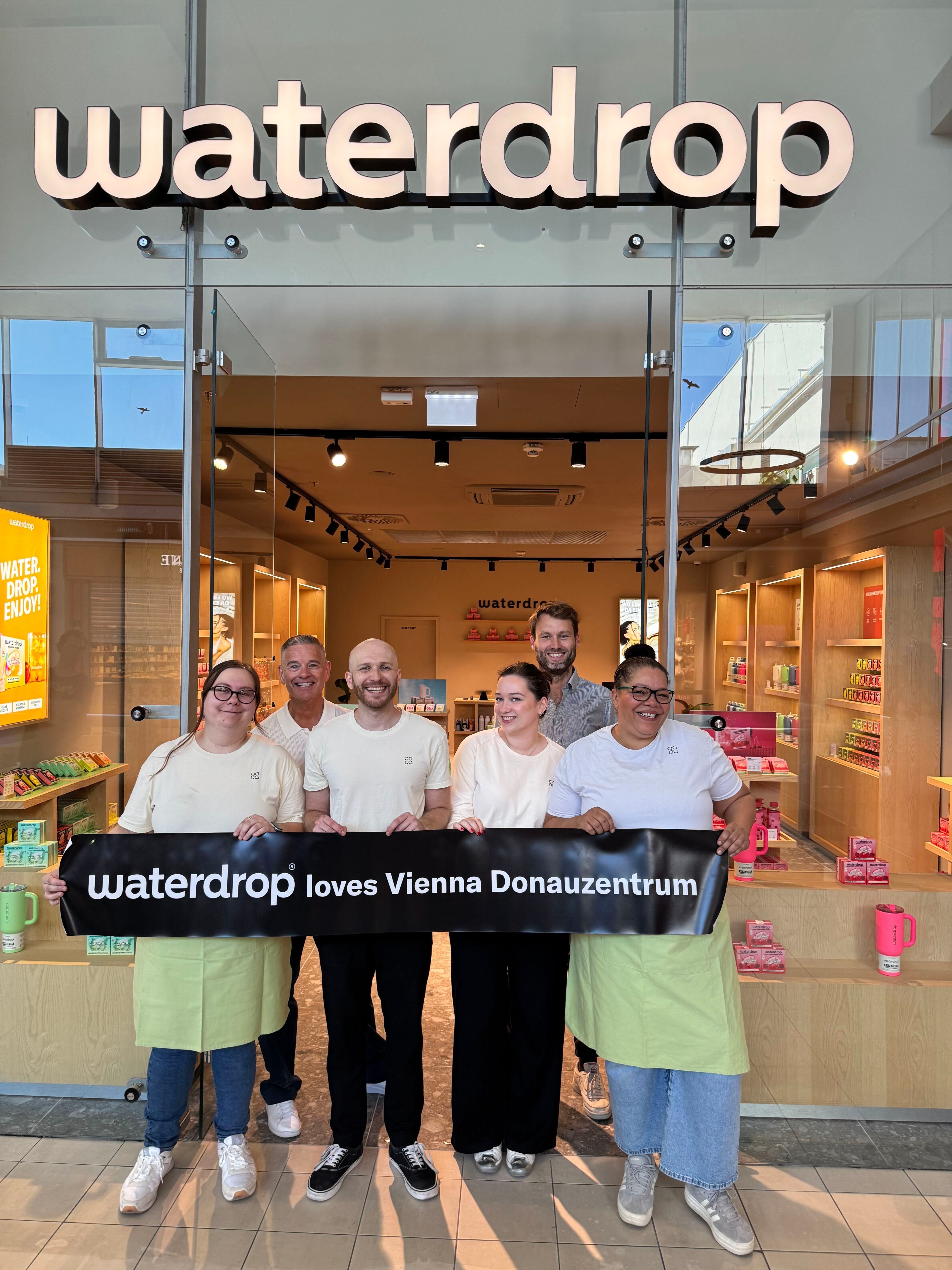 Sechs Personen stehen lachend vor dem Waterdrop-Store im Donauzentrum mit einem Banner.