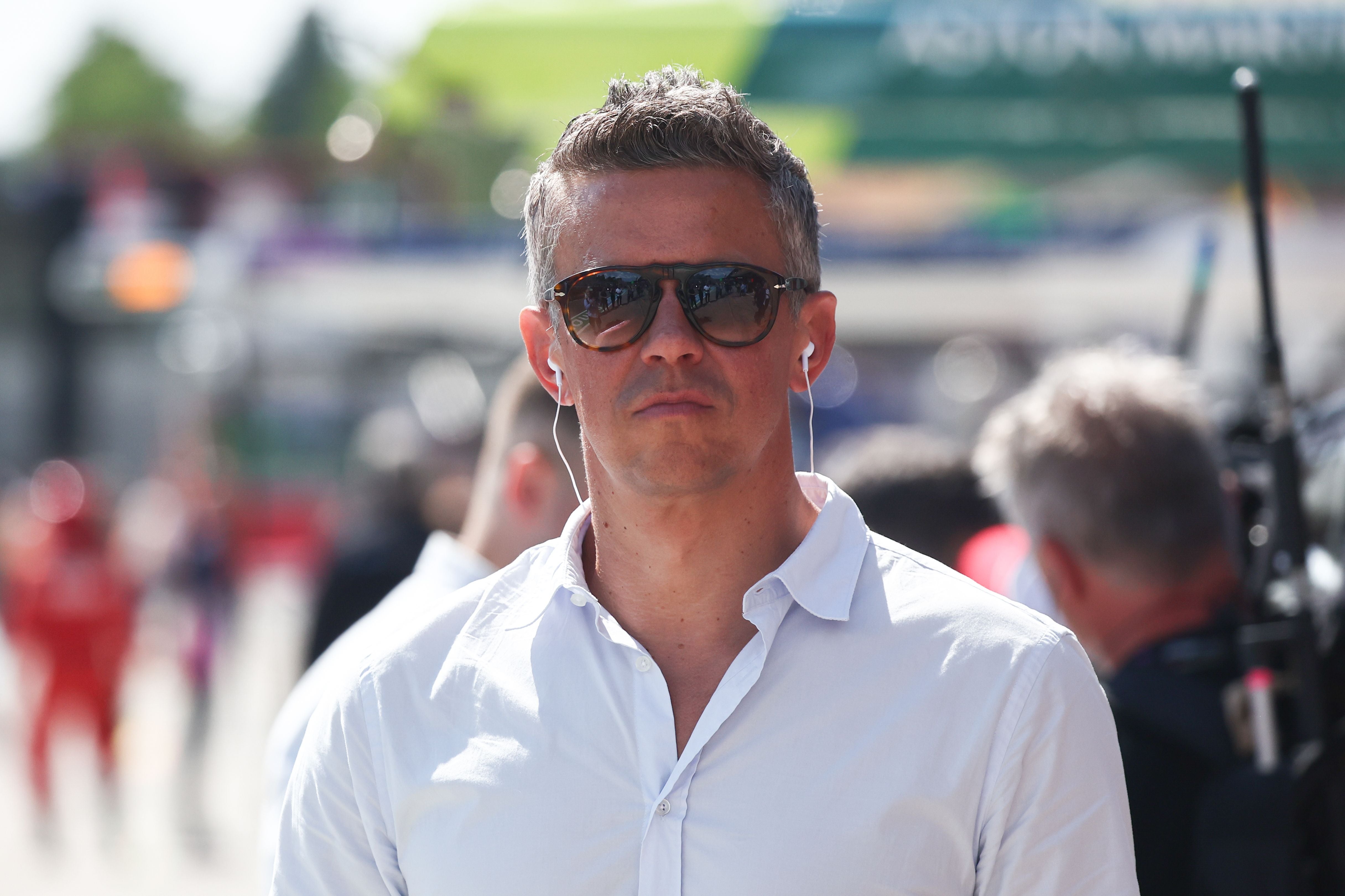 Ein Mann mit Sonnenbrille und Kopfhörern trägt ein weißes Hemd auf einer Rennstrecke.