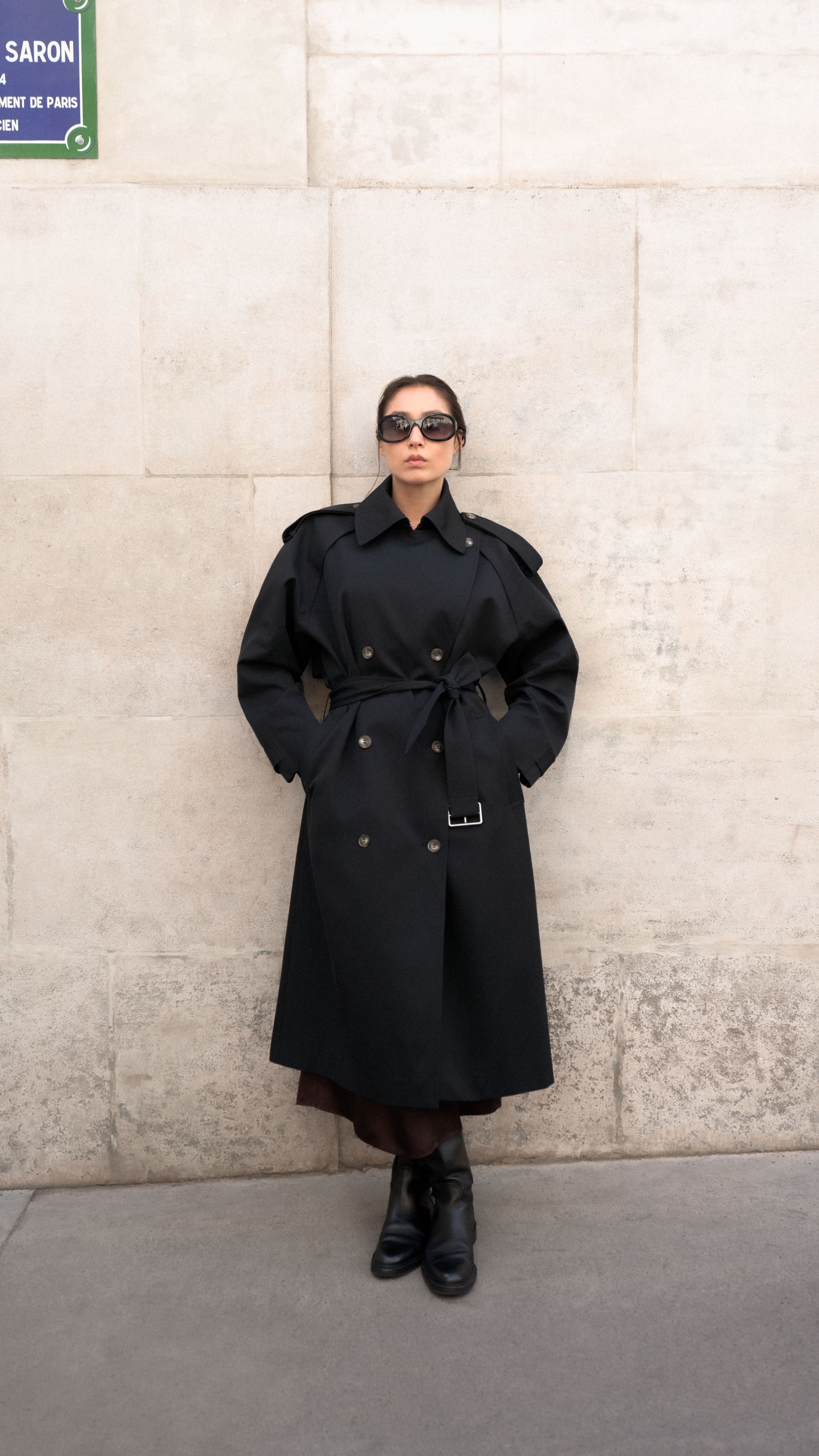 Frau in schwarzem Trenchcoat, Sonnenbrille und Stiefeln steht vor einer Steinwand.