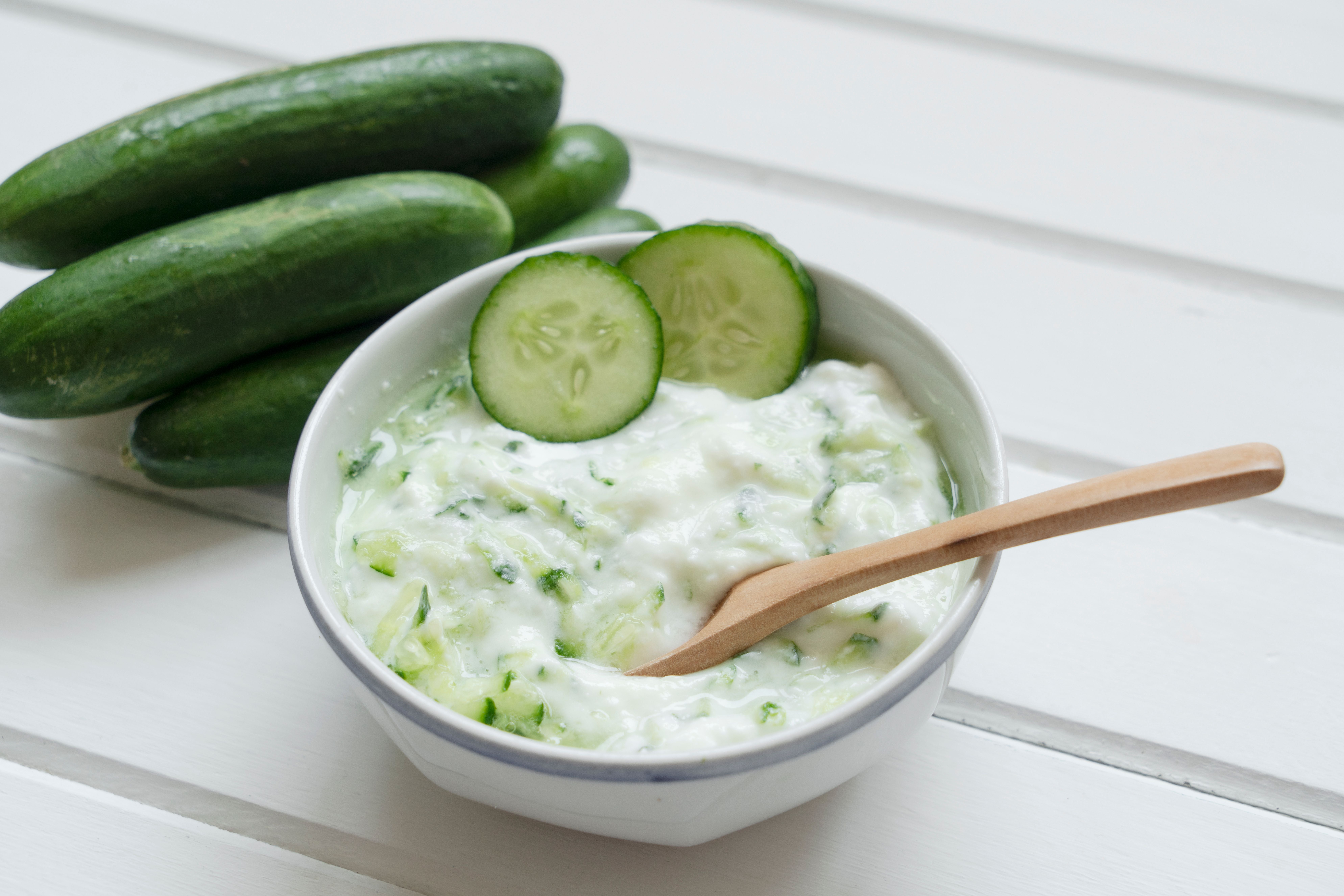Schale mit Tzatziki und Holzlöffel, daneben liegen frische Gurken auf einem weißen Tisch.
