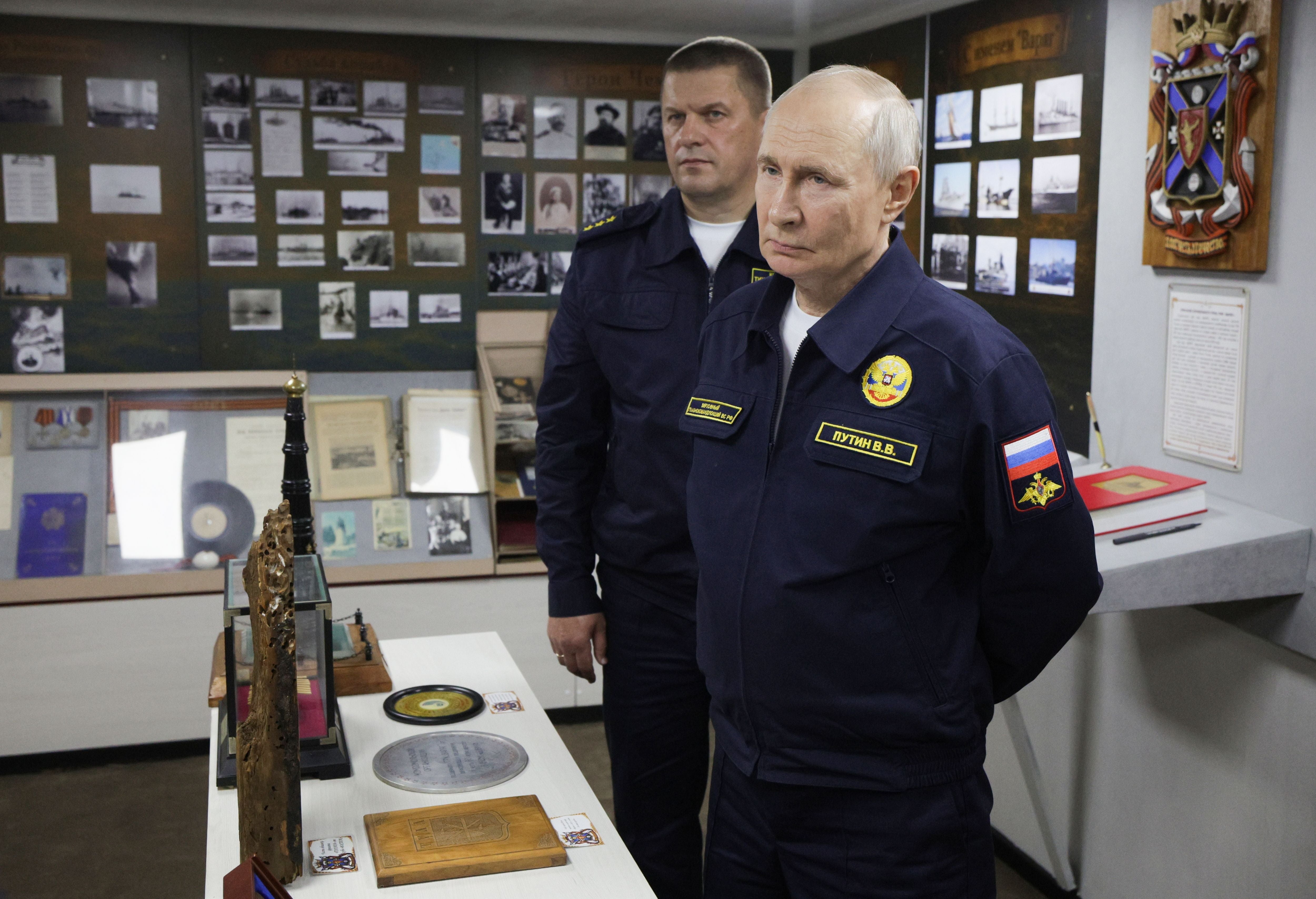 Zwei Männer in Marineuniform stehen in einem Ausstellungsraum mit Fotografien und Andenken.