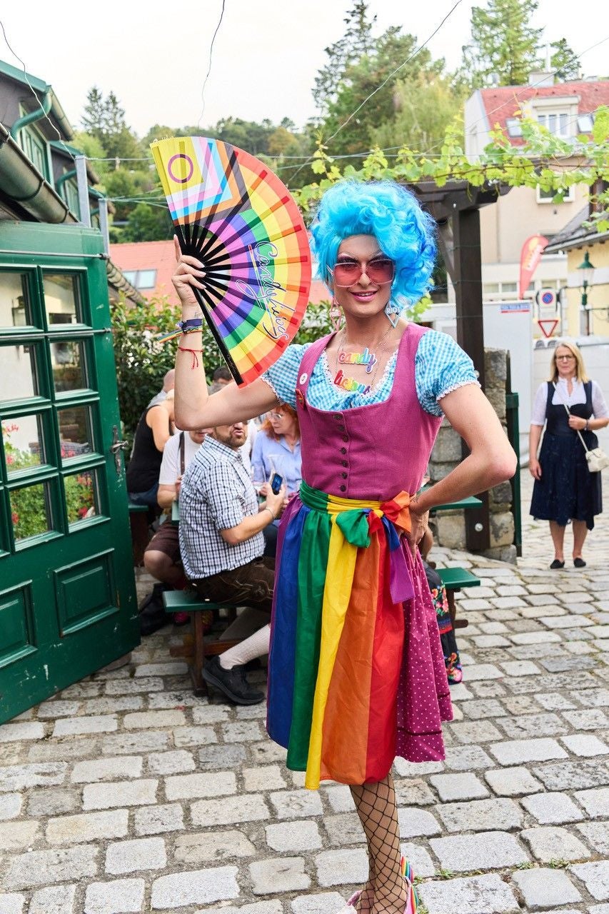 Dragqueen mit blauer Perücke und Regenbogenrock beim Neustifter Kirtag, hält bunten Fächer.