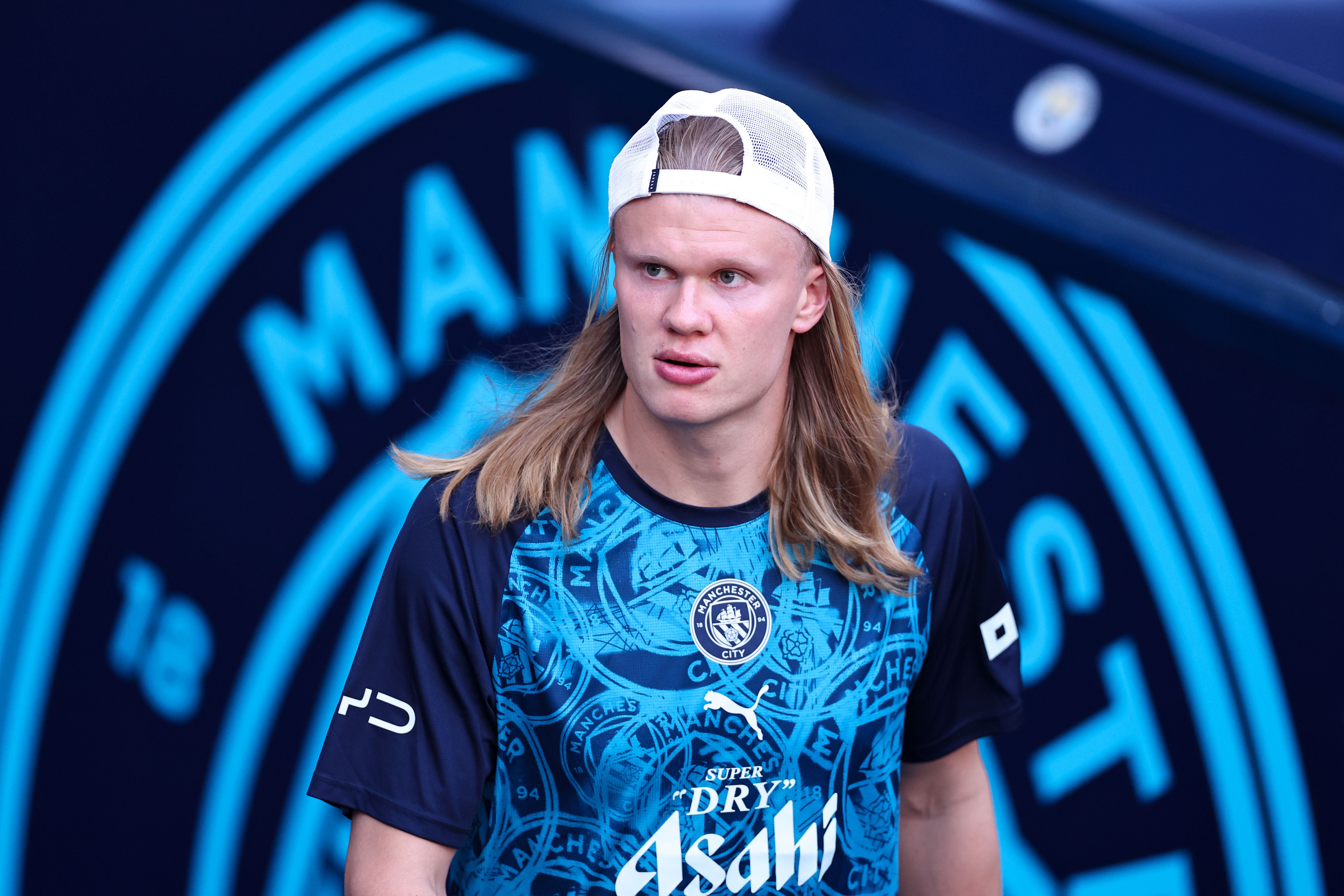 Ein Fußballspieler im Manchester City-Trikot steht vor einem Vereinslogo und trägt eine weiße Kappe.