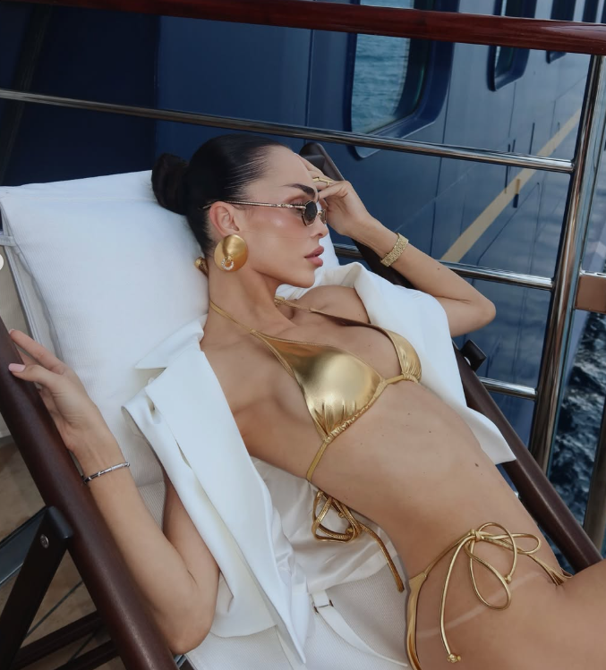 Frau im goldenen Bikini und weißem Blazer entspannt auf einer Yacht-Liege.
