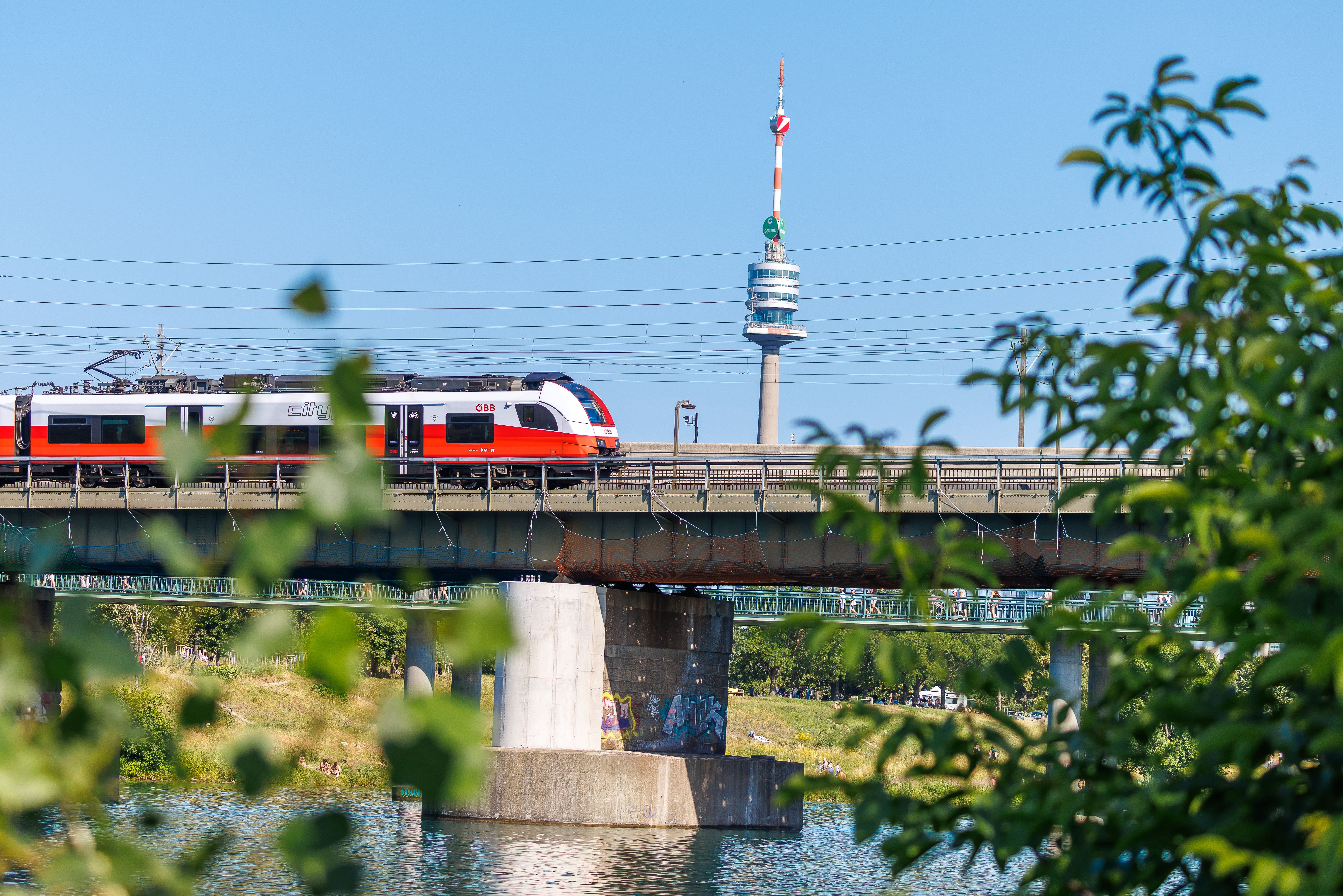 Eine ÖBB-Schnellbahn fährt über eine Brücke, im Hintergrund der Wiener Donauturm bei blauem Himmel.