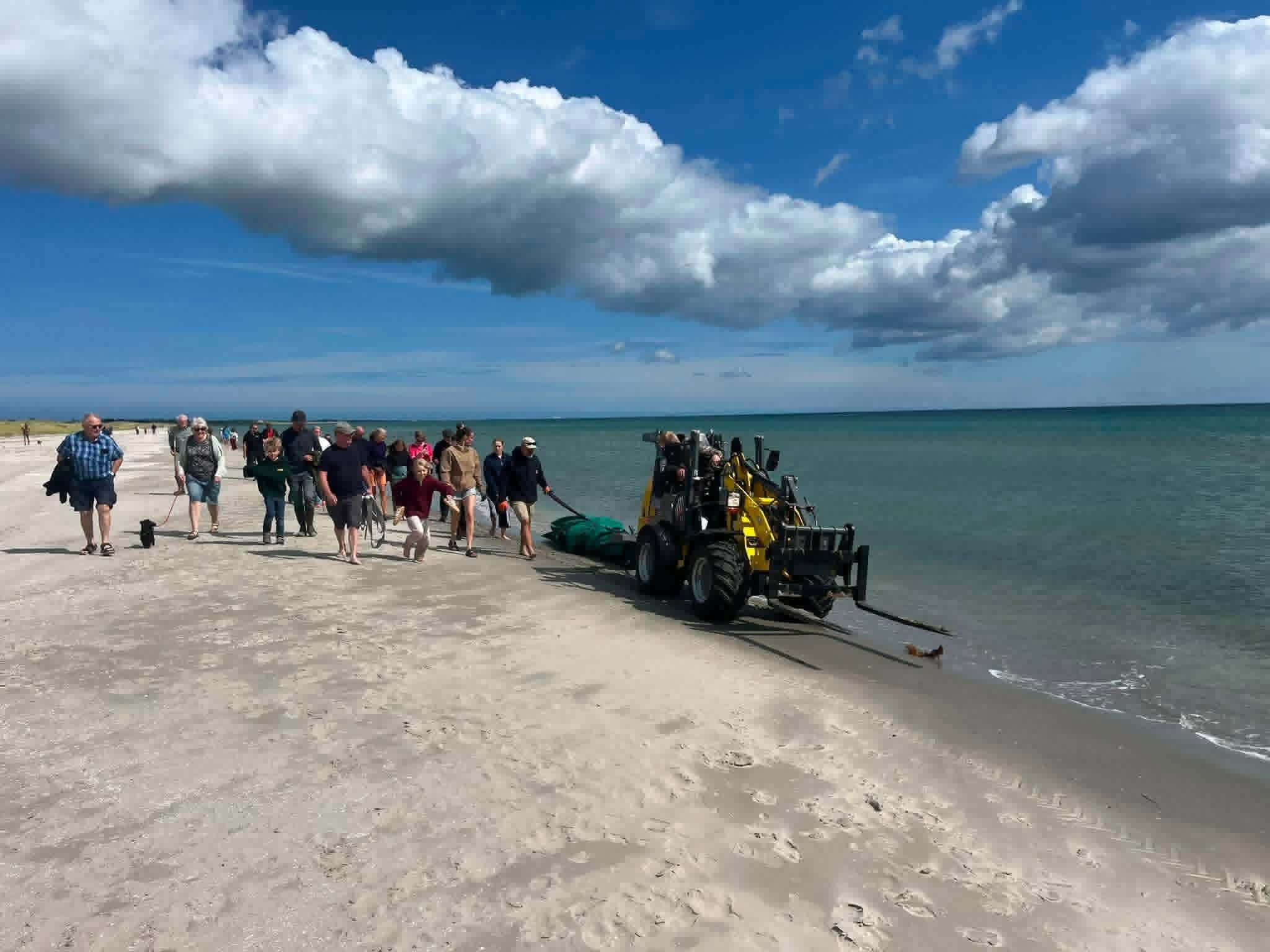 Mehrere Personen und ein Bagger am Sandstrand neben der Nordsee unter blauem Himmel.