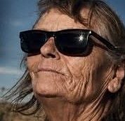 Ältere Frau mit Sonnenbrille blickt in die Ferne, Hintergrund ist wolkiger Himmel.