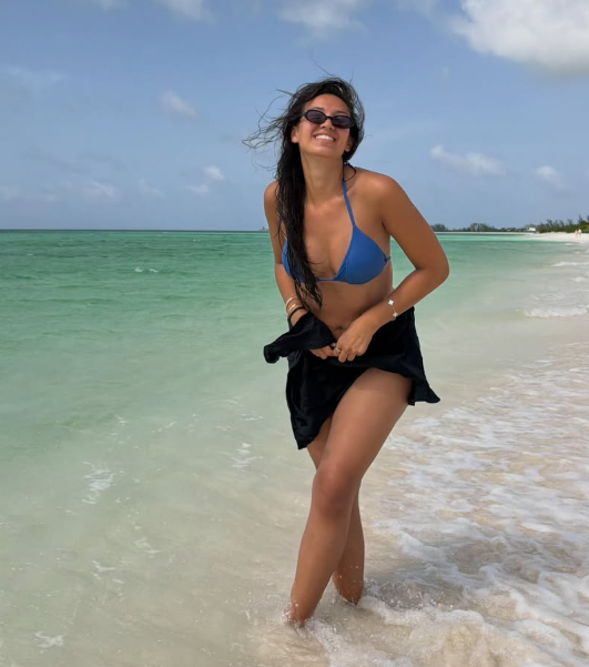 Frau im blauen Bikini lächelt am Strand mit türkisfarbenem Meer im Hintergrund.
