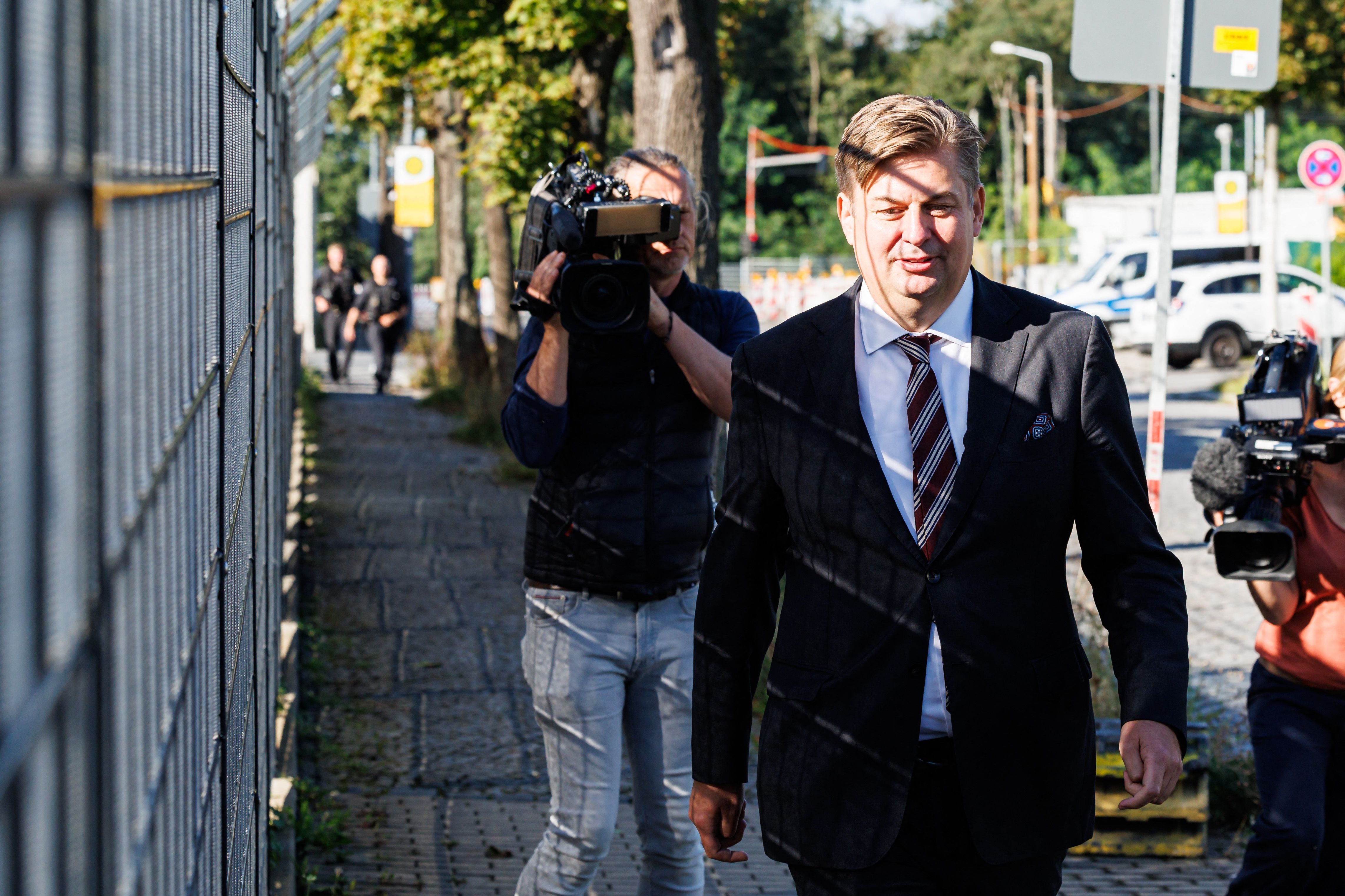 Ein Mann im Anzug geht an Kamerateams vorbei auf einem Gehweg an einem Zaun entlang.