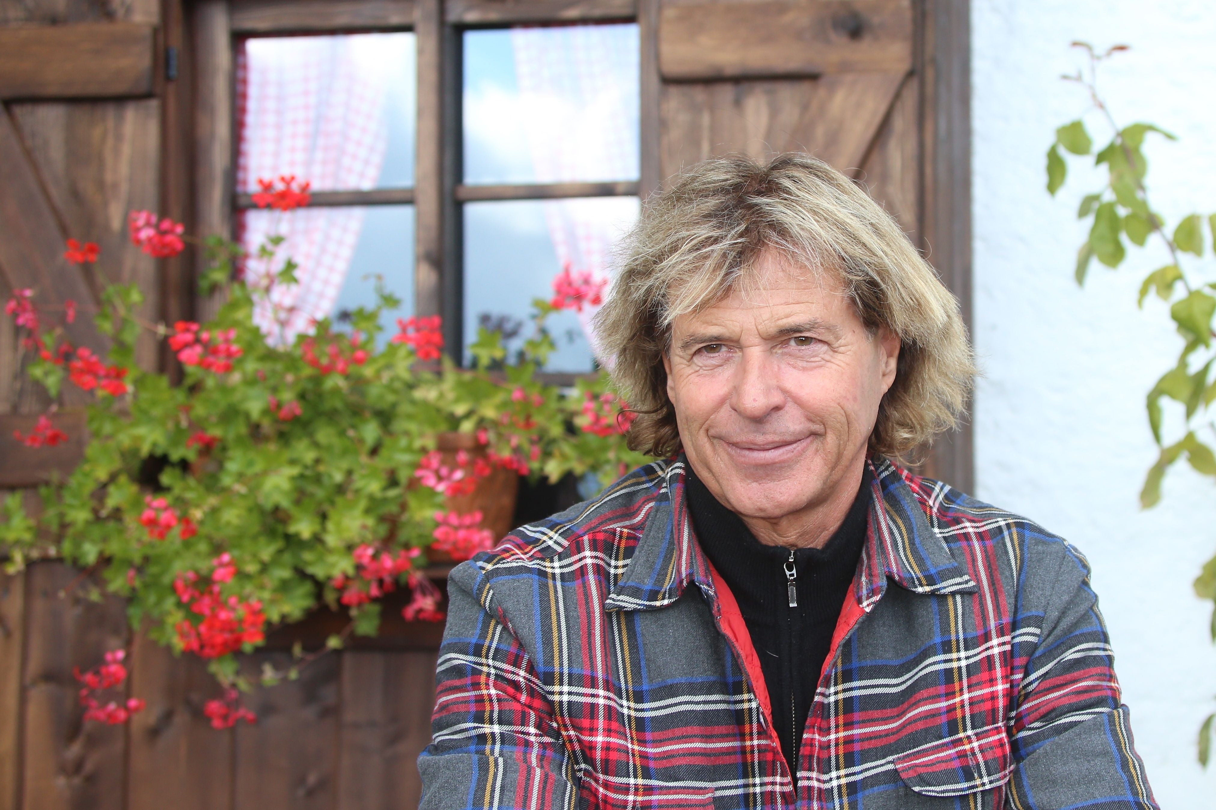 Ein Mann mit mittellangem blondem Haar sitzt vor einem Fenster mit roten Blumen.