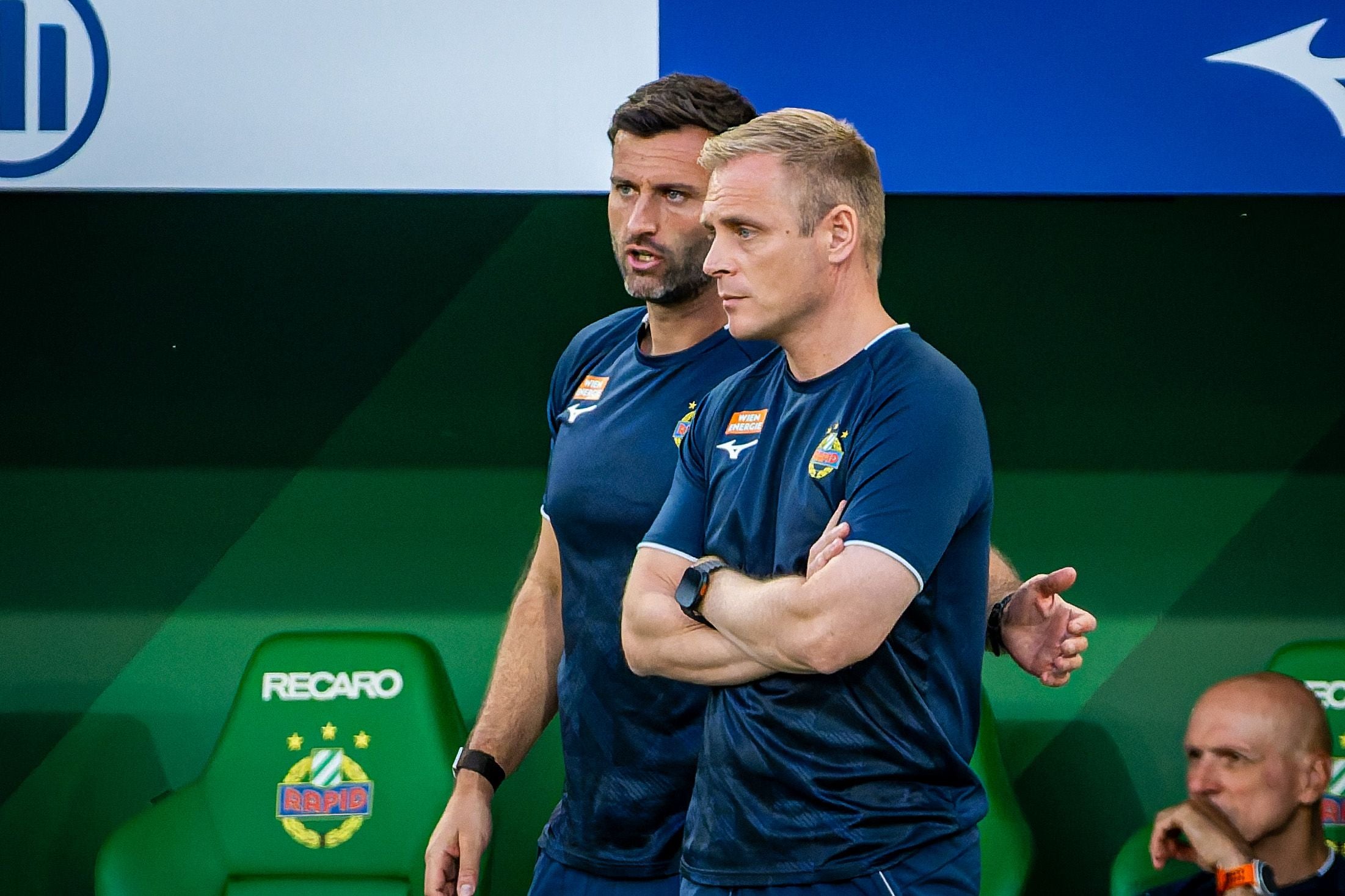 Zwei Rapid-Trainer im Gespräch vor grüner Sitzbank im Fußballstadion.