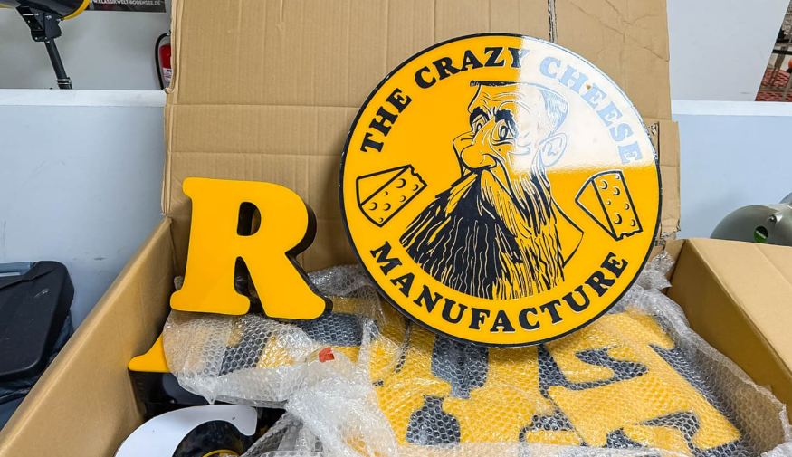 Gelbes Schild mit "The Crazy Cheese Manufacture" und gezeichnetem bärtigem Gesicht in Karton.