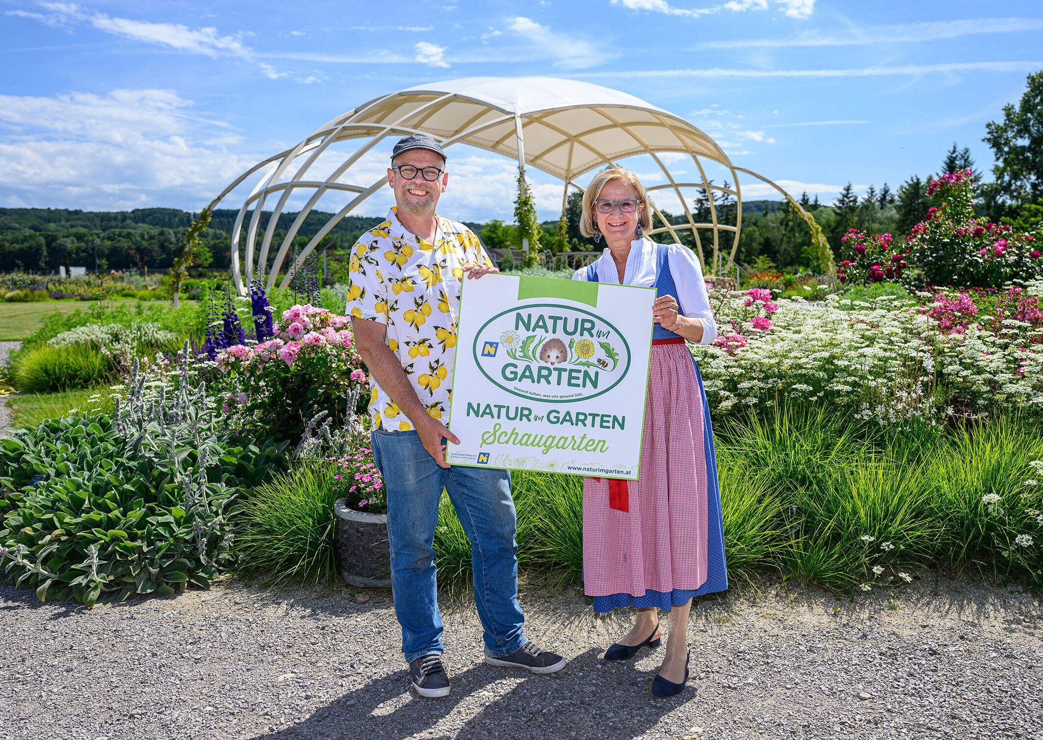 Ein Mann und eine Frau halten ein "Natur im Garten"-Schild in einem blühenden Schaugarten.