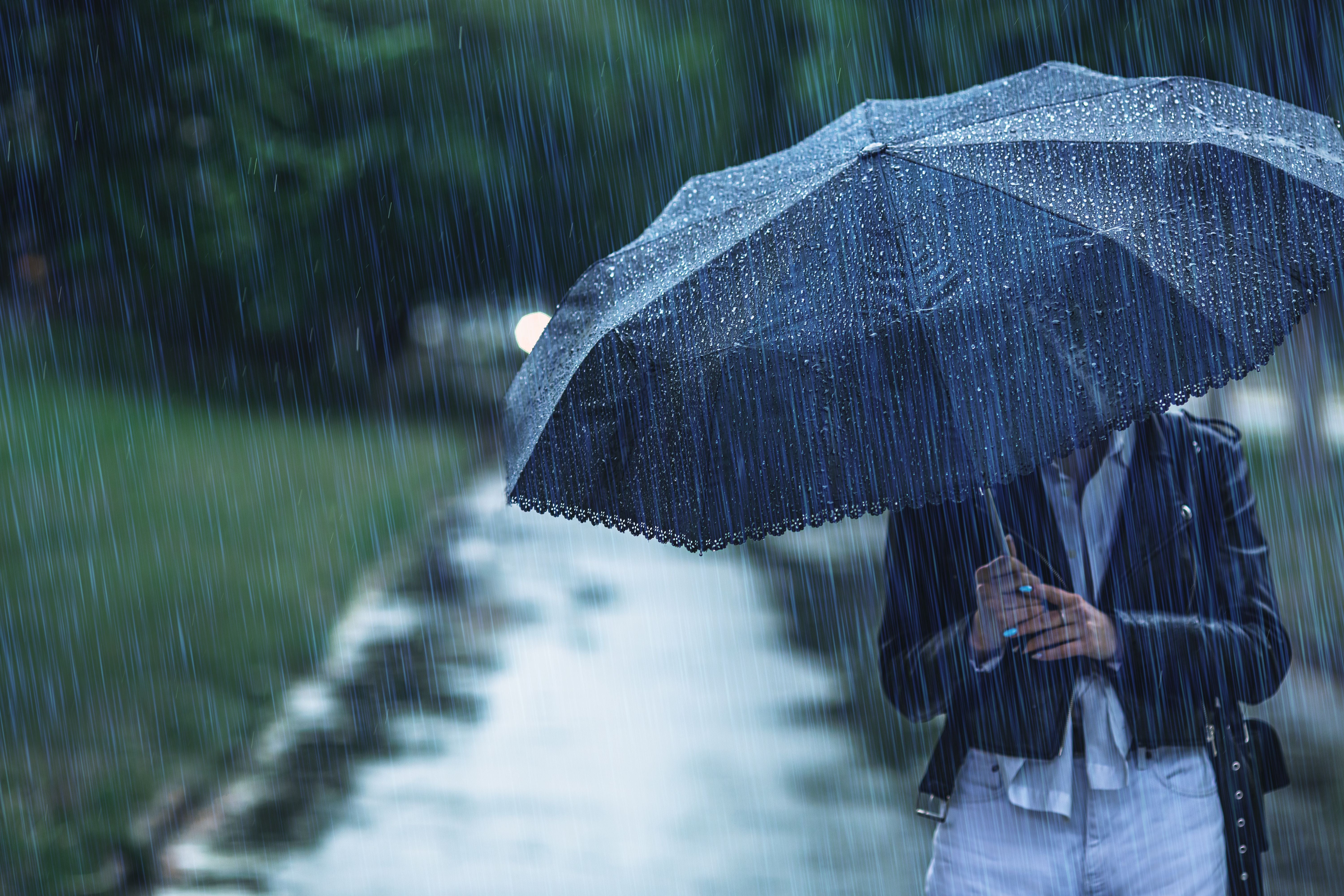 Person mit schwarzem Regenschirm steht im Regen auf einem nassen Gehweg.