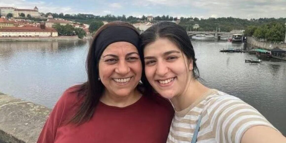 Zwei Frauen lächeln für ein Selfie vor einem Fluss mit Brücke und Stadt im Hintergrund.