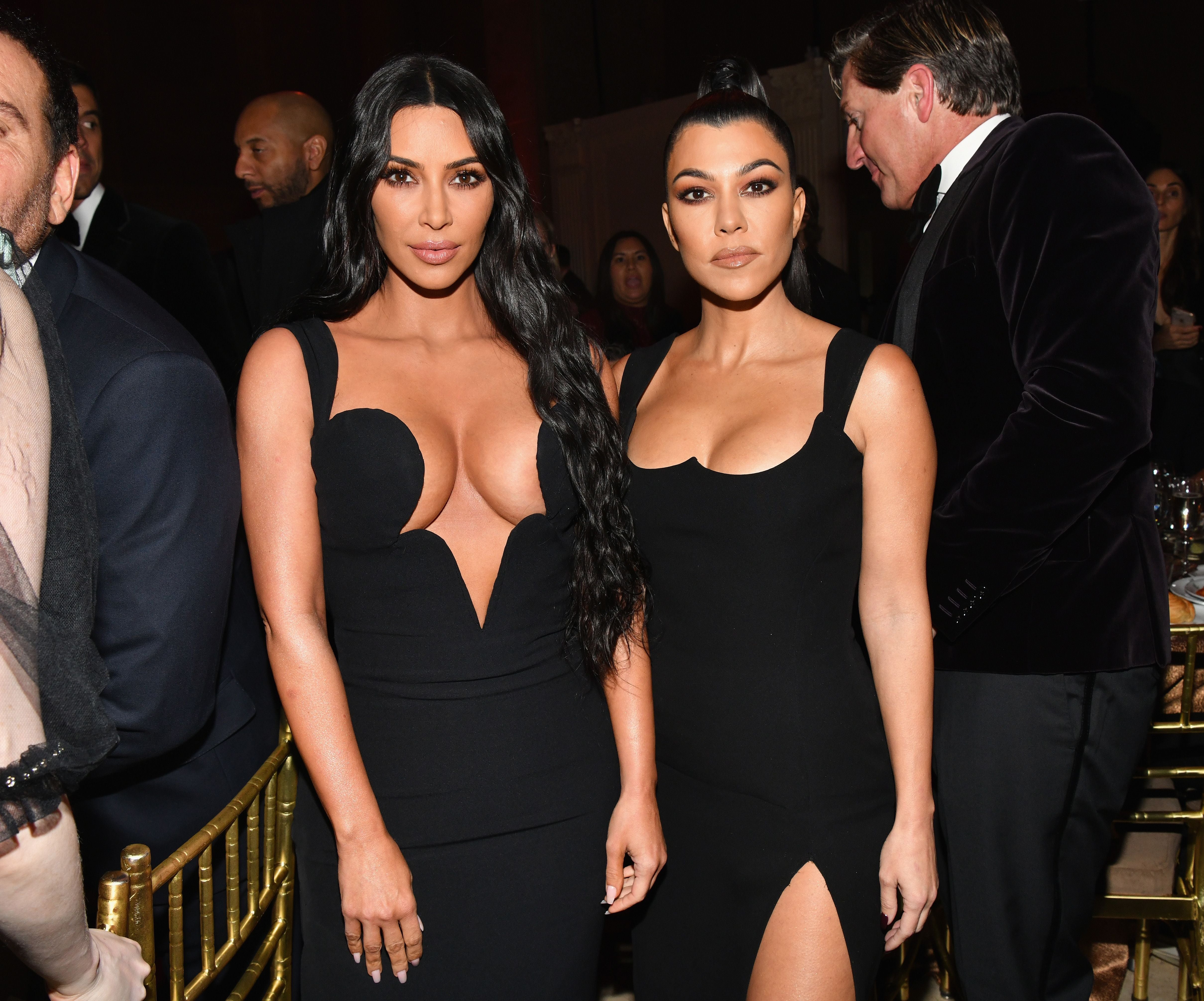 Zwei Frauen in eleganten schwarzen Kleidern posieren auf einer Gala mit weiteren Gästen im Hintergrund.
