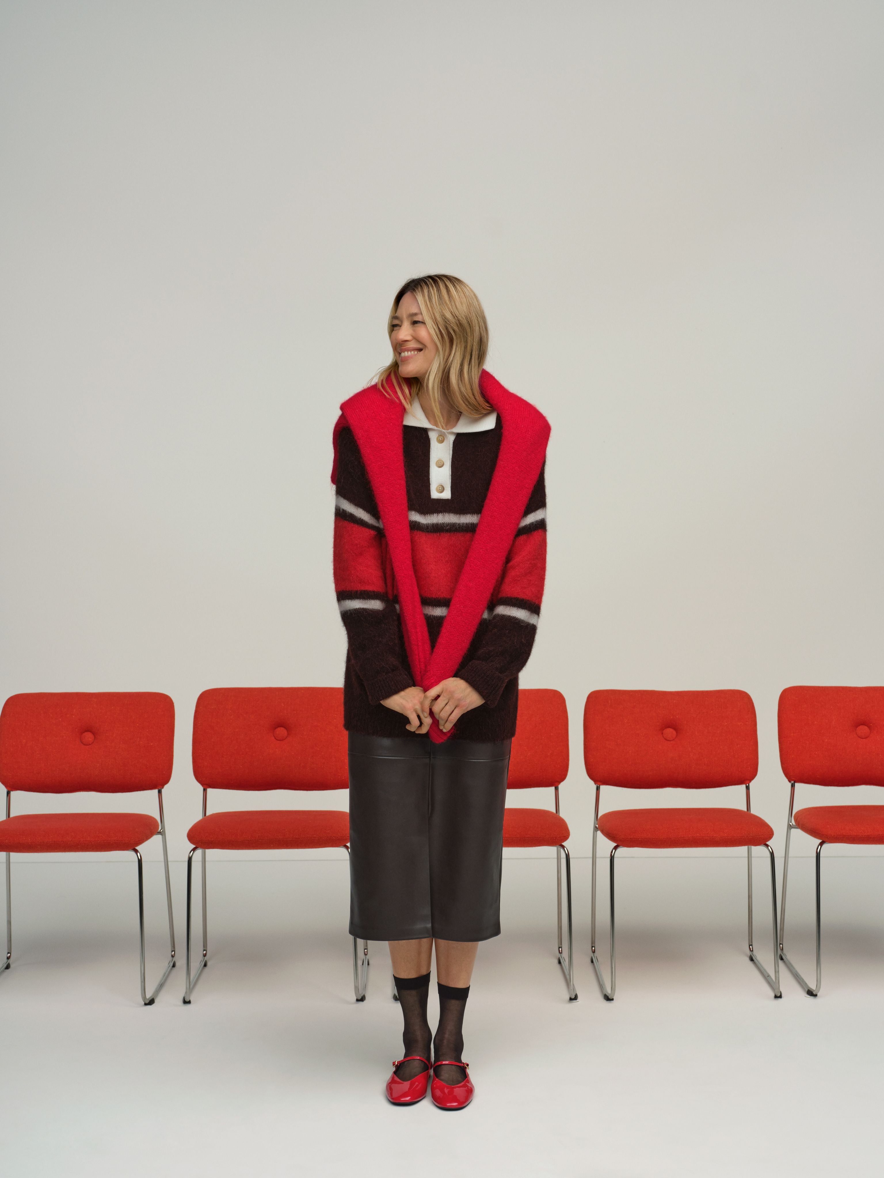 Frau in rotem Pullover und schwarzem Rock steht lächelnd vor einer Reihe roter Stühle.