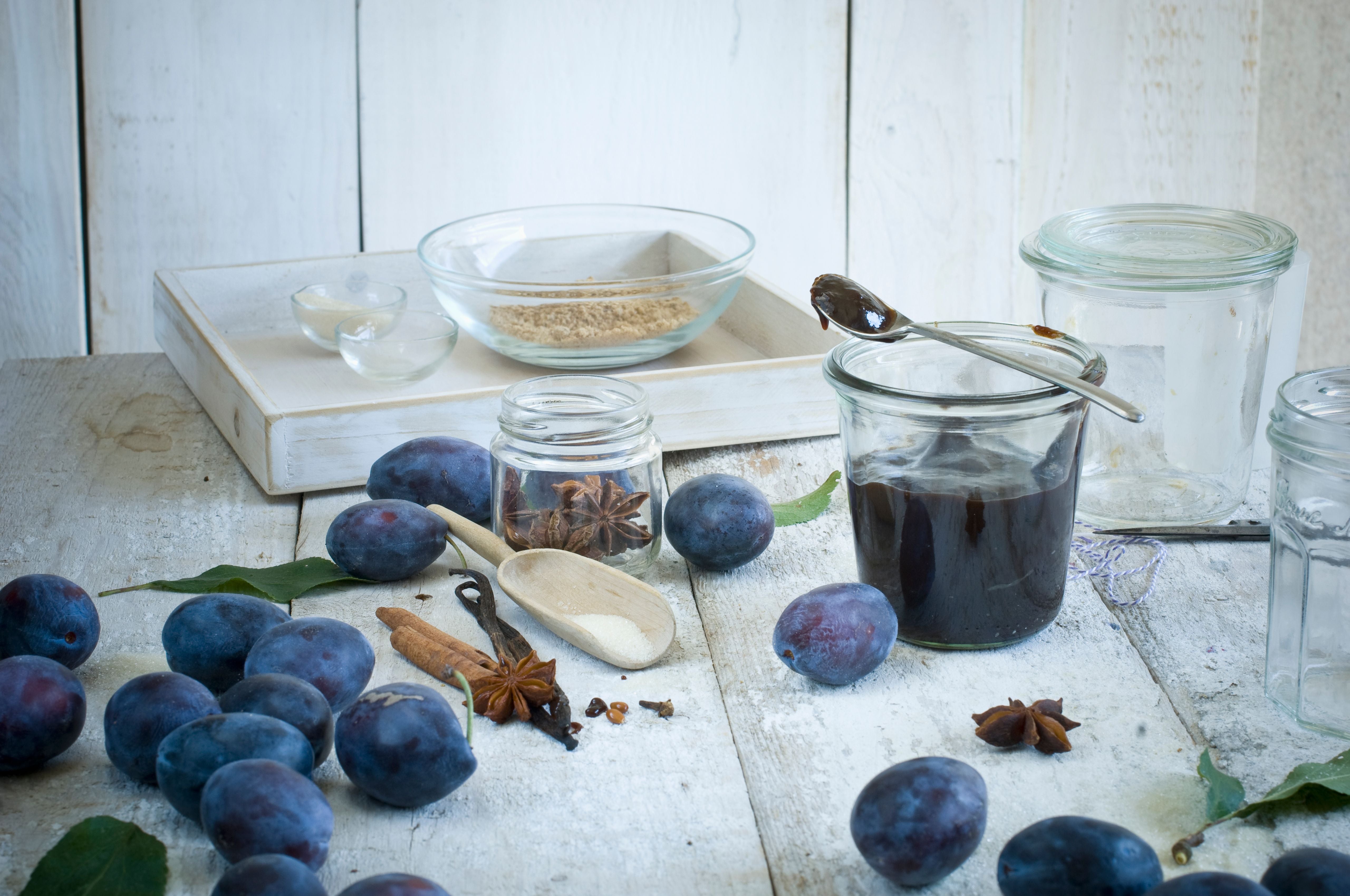 Zwetschgen, Gewürze und ein Glas Pflaumenmus auf einem hellen Holztisch arrangiert.