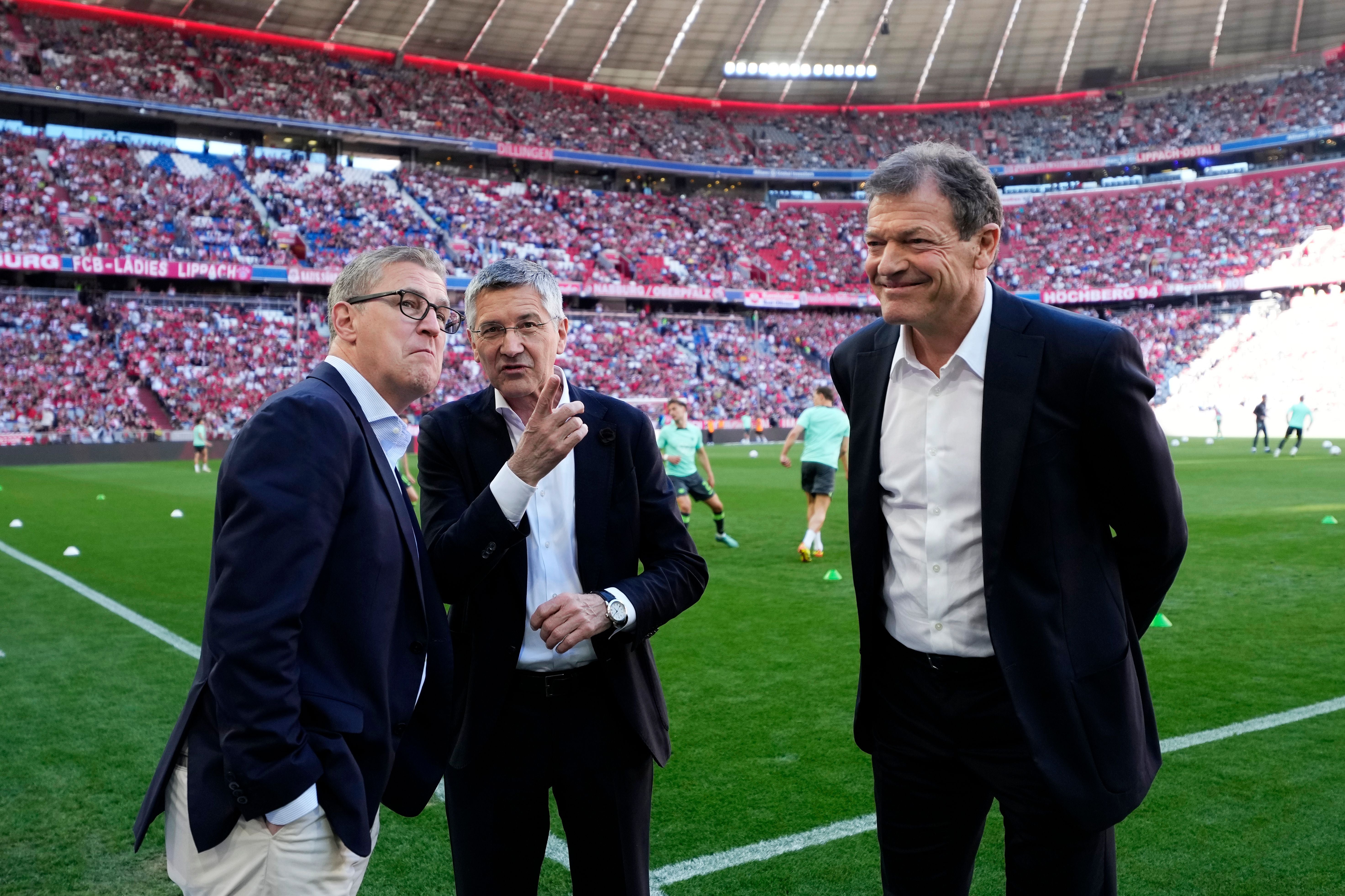 Drei Männer im Anzug unterhalten sich am Spielfeldrand in einem vollen Fußballstadion.