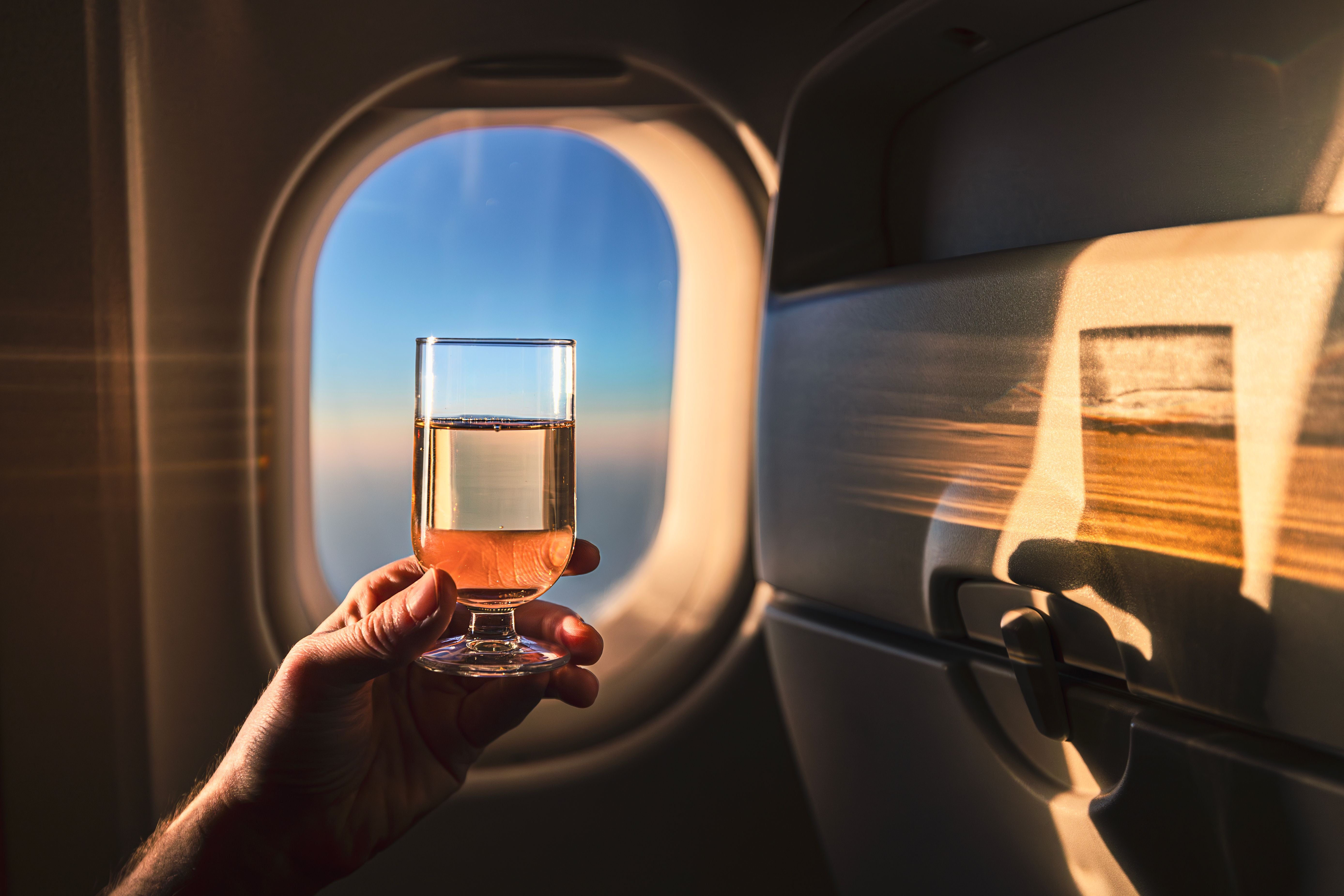 Hand hält ein Champagnerglas vor einem Flugzeugfenster bei Sonnenaufgang.