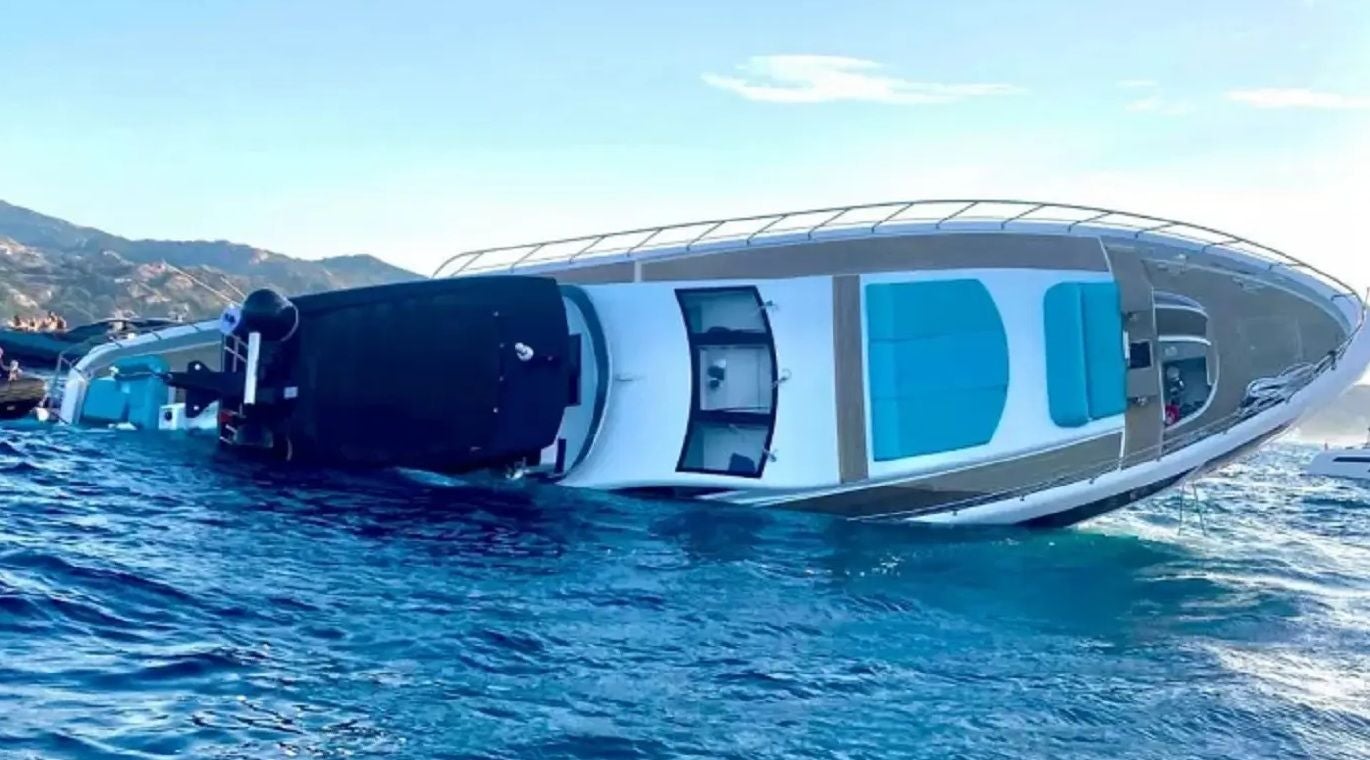 Eine gekippte Jacht droht im Meer zu sinken, Berge im Hintergrund.