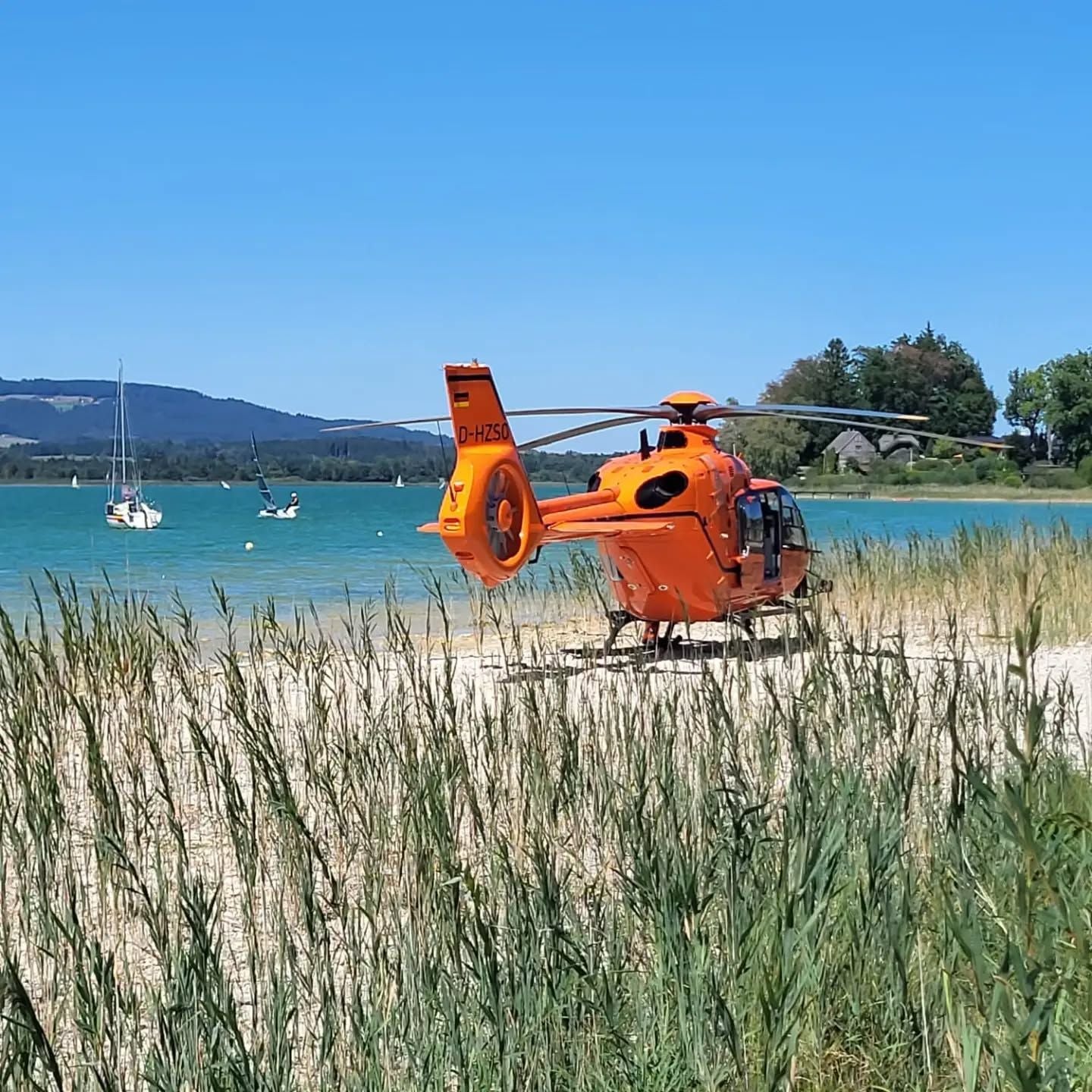 Ein orangefarbener Rettungshubschrauber steht am Ufer eines Sees, im Hintergrund Segelboote.