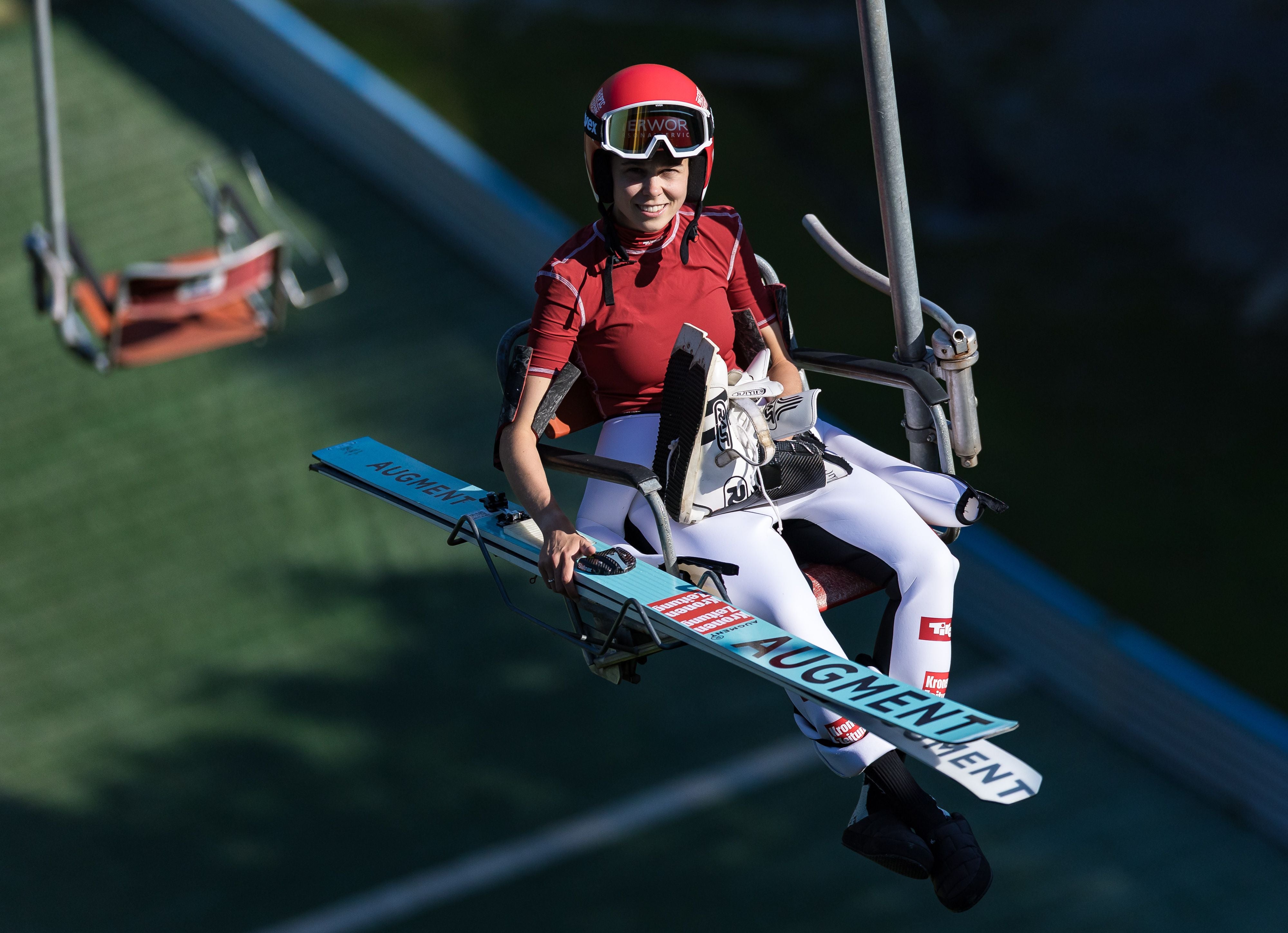 Skispringerin Lisa Eder sitzt mit Ausrüstung auf einem Sessellift und lächelt in die Kamera.
