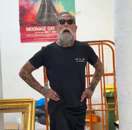 Ein tätowierter Mann mit Sonnenbrille steht mit den Händen in der Hüfte in einem Atelier.