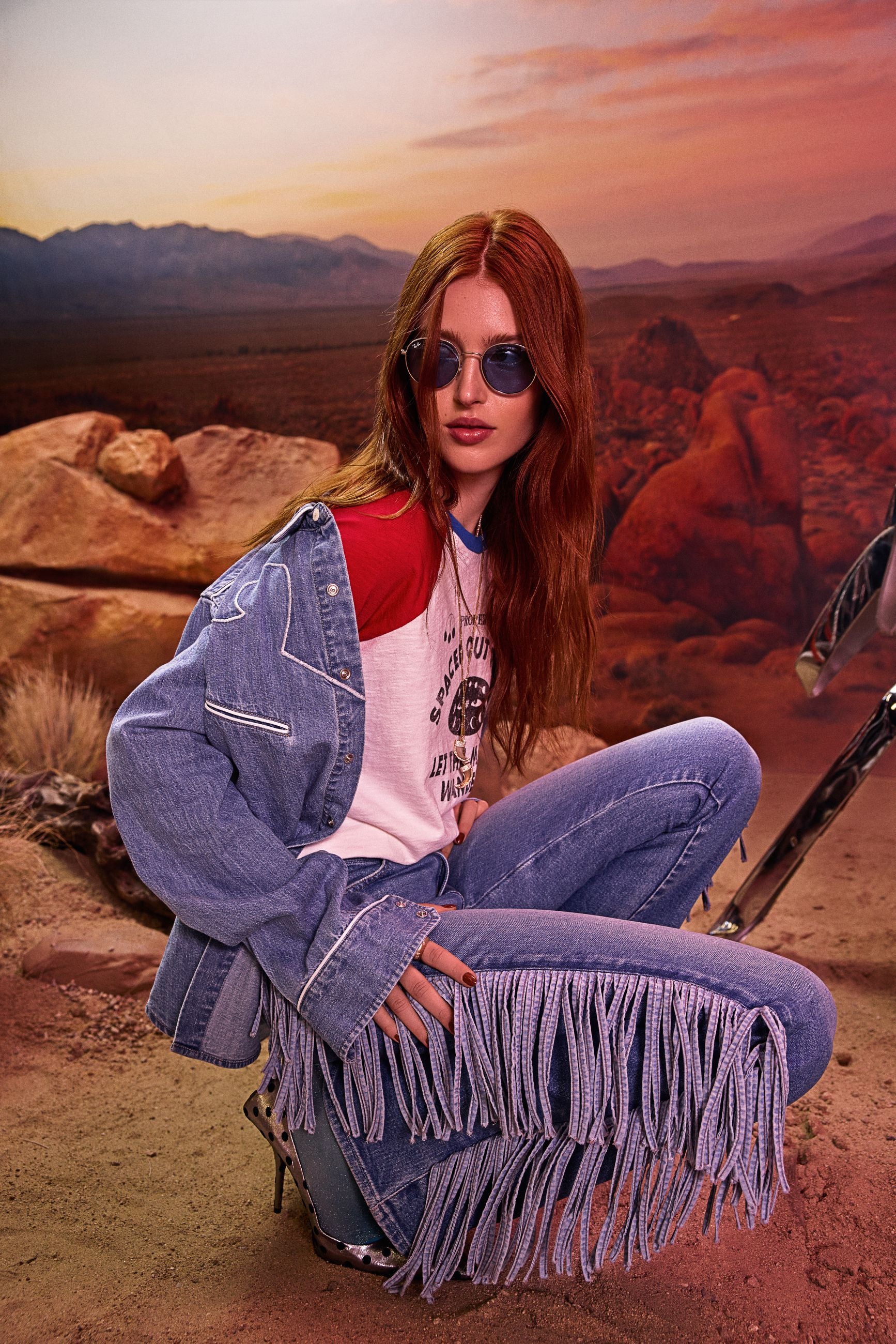 Frau mit Sonnenbrille und Jeans mit Fransen sitzt in wüstenähnlicher Landschaft bei Sonnenuntergang.