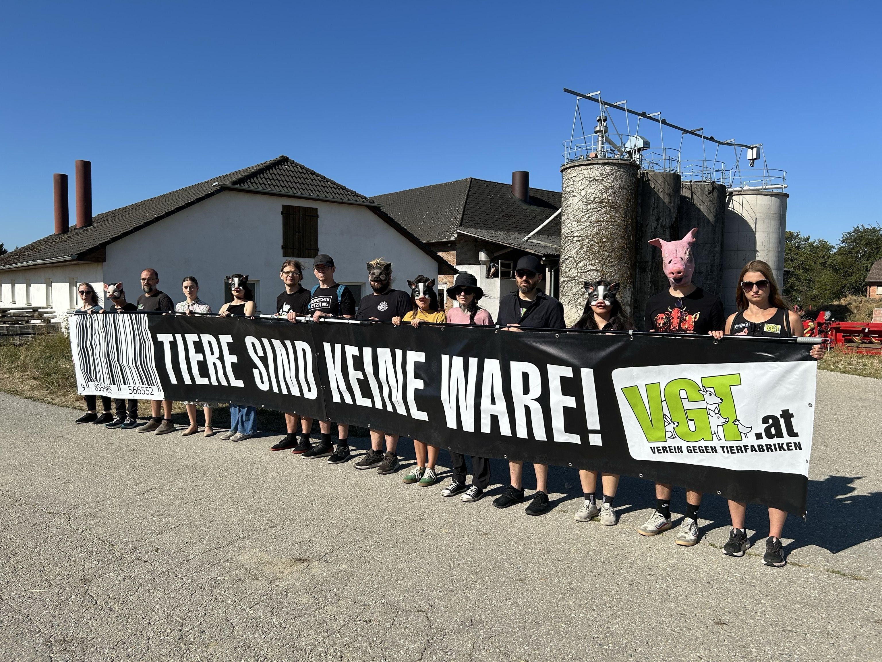 Menschen mit Masken halten ein Banner mit der Aufschrift „Tiere sind keine Ware!“ vor einem Bauernhof.