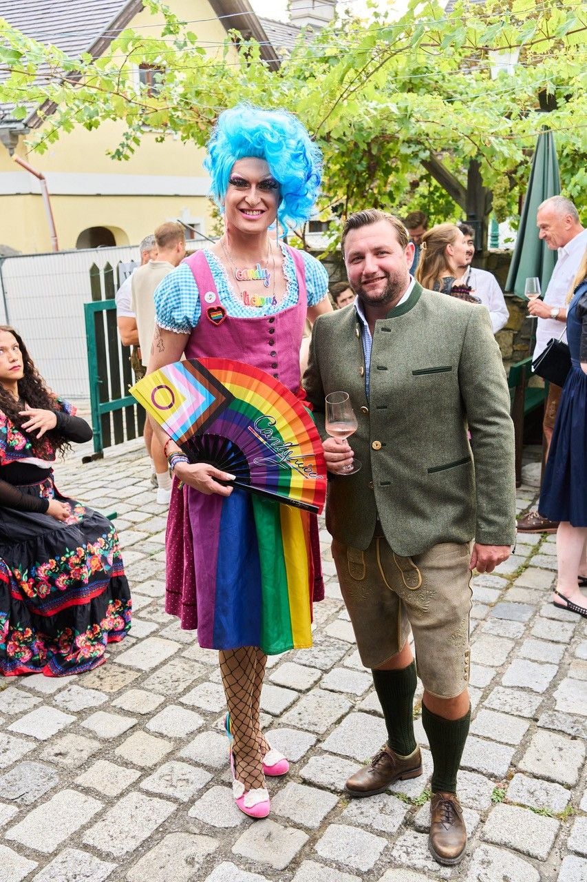 Drag Queen mit blauer Perücke und Regenbogenfächer steht neben Mann in Tracht beim Neustifter Kirtag.