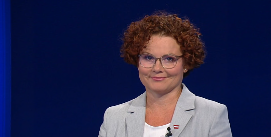 Eine Frau mit lockigen Haaren, Brille und grauem Blazer vor blauem Hintergrund.