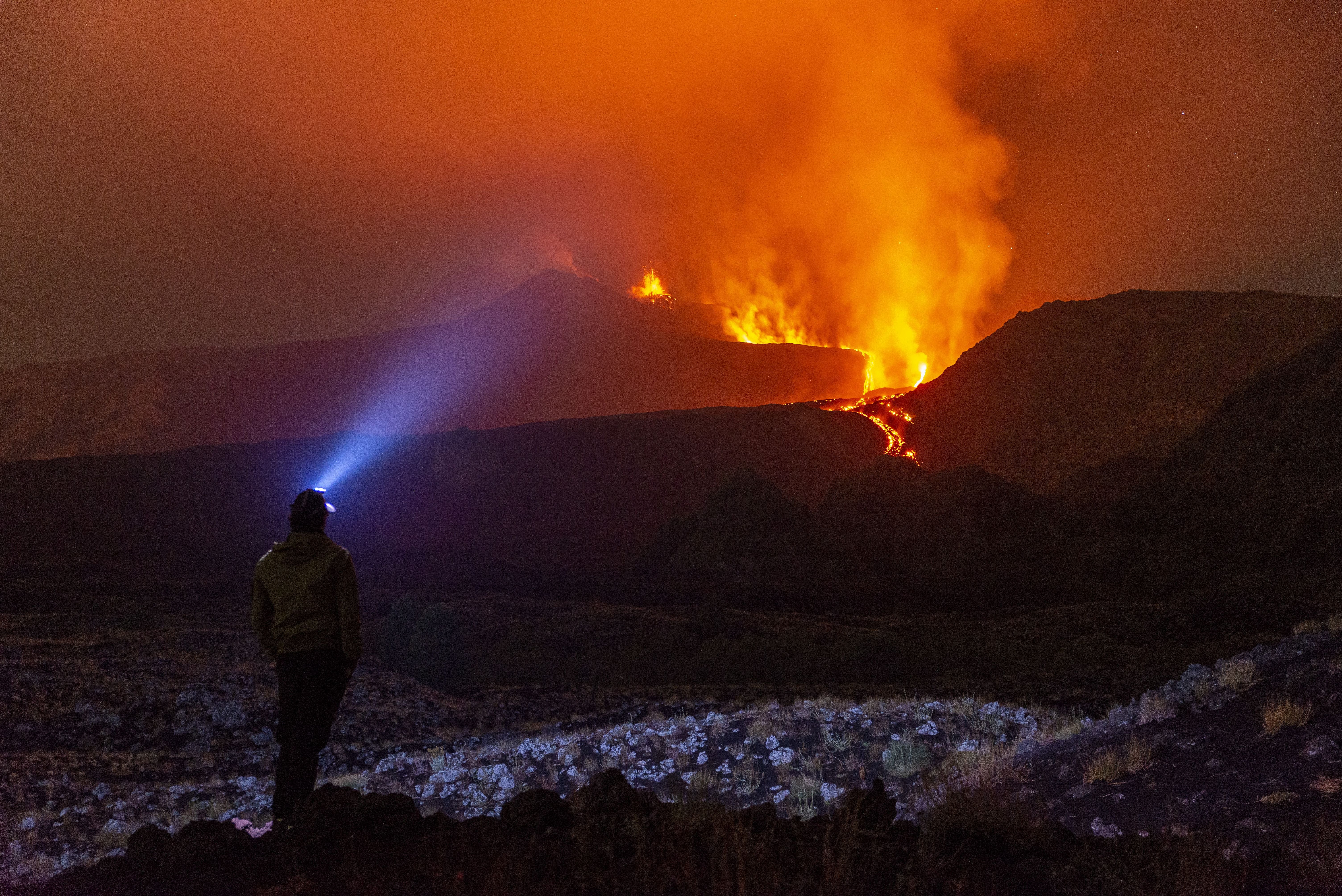 Person mit Stirnlampe beobachtet nachts einen ausbrechenden Vulkan mit glühender Lava und Aschewolken.