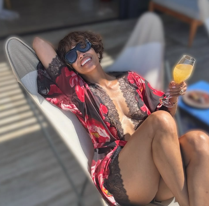 Frau in Sonnenbrille relaxt auf einer Liege mit einem Glas Orangensaft und lächelt in die Sonne.