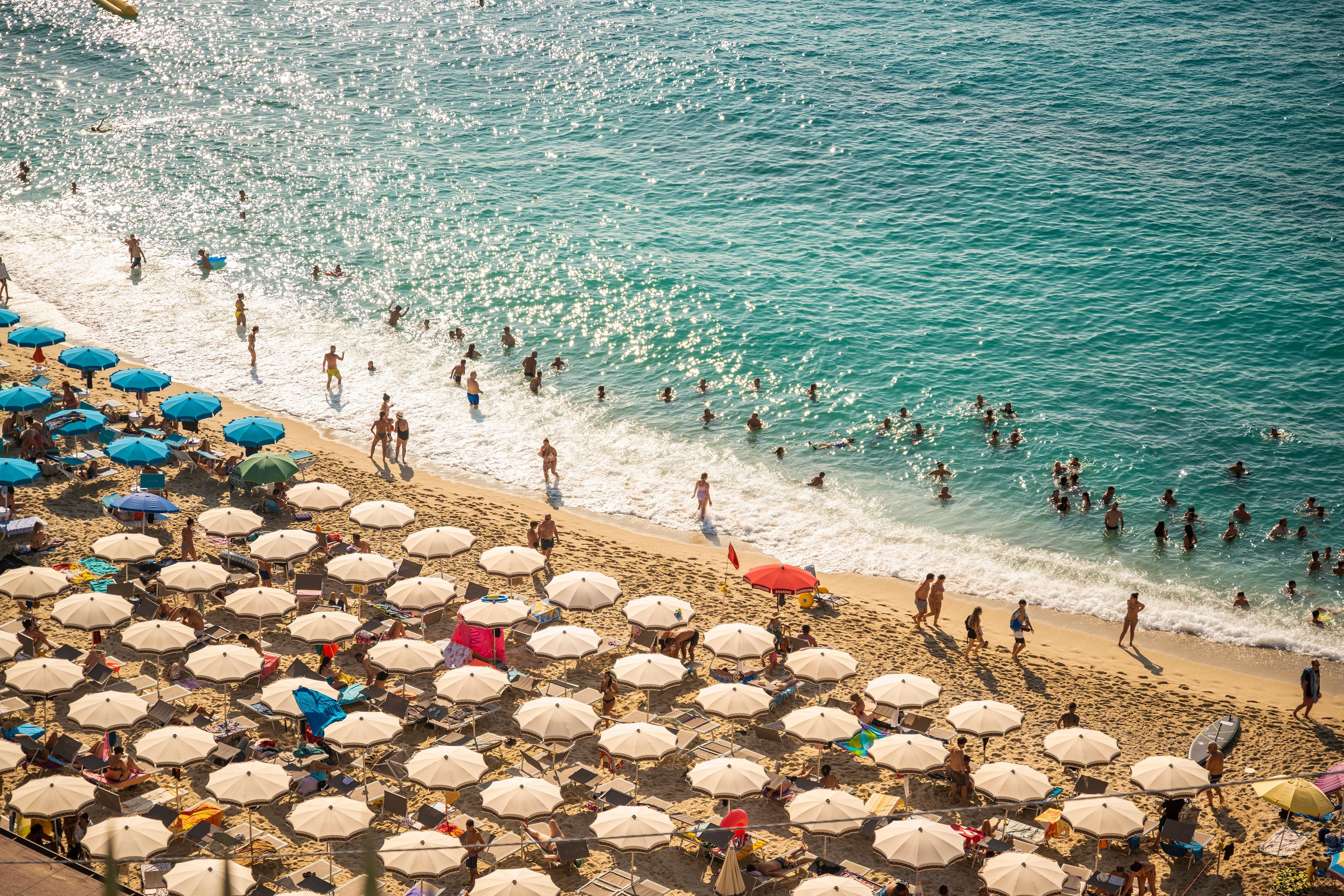 Überfüllter Sandstrand in Italien mit vielen Sonnenschirmen und schwimmenden Menschen im Meer.