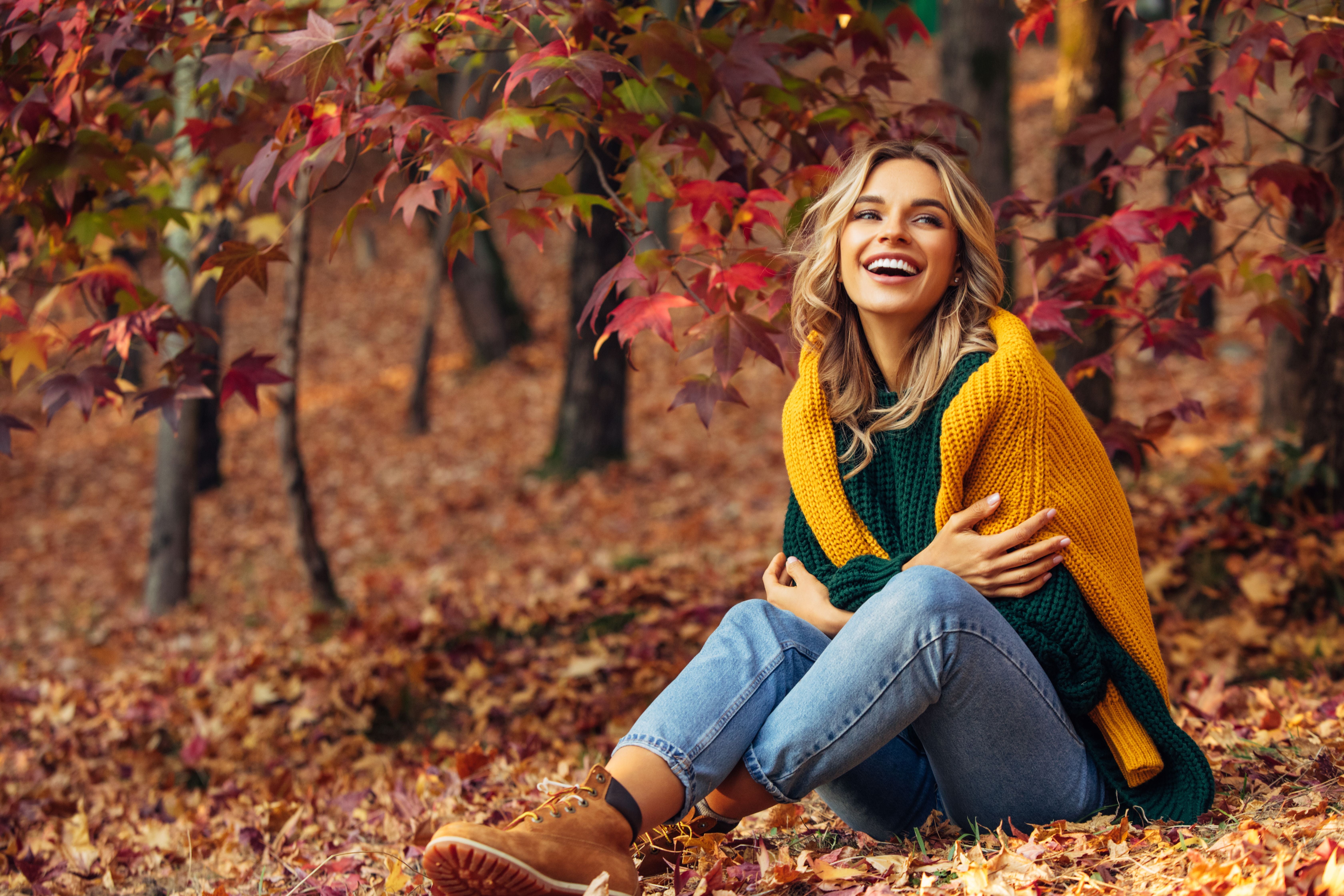 Junge Frau sitzt lachend im Herbstlaub unter bunten Bäumen und trägt einen gelben Schal.