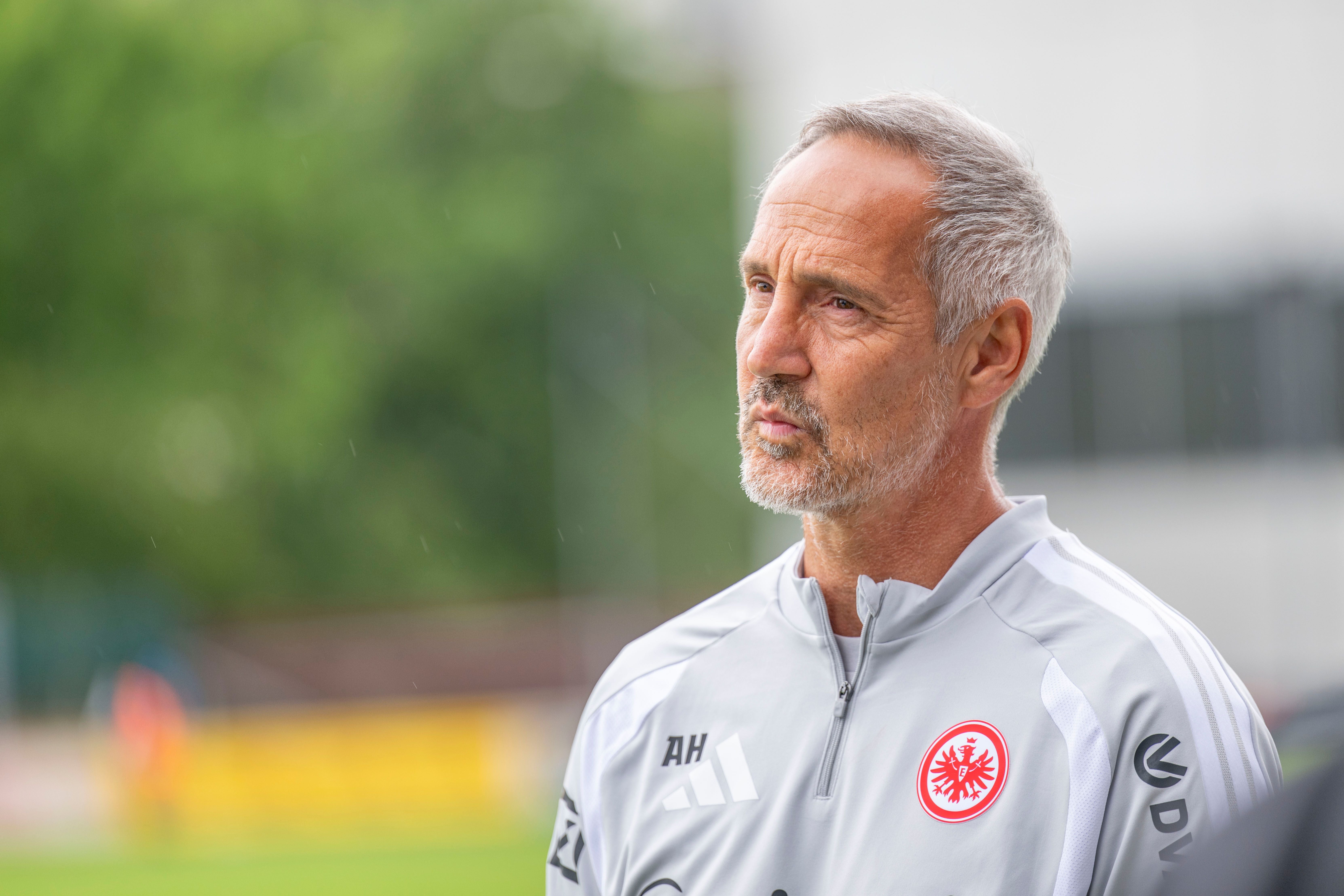 Ein Fußballtrainer im grauen Eintracht-Frankfurt-Trainingsanzug steht nachdenklich im Freien.