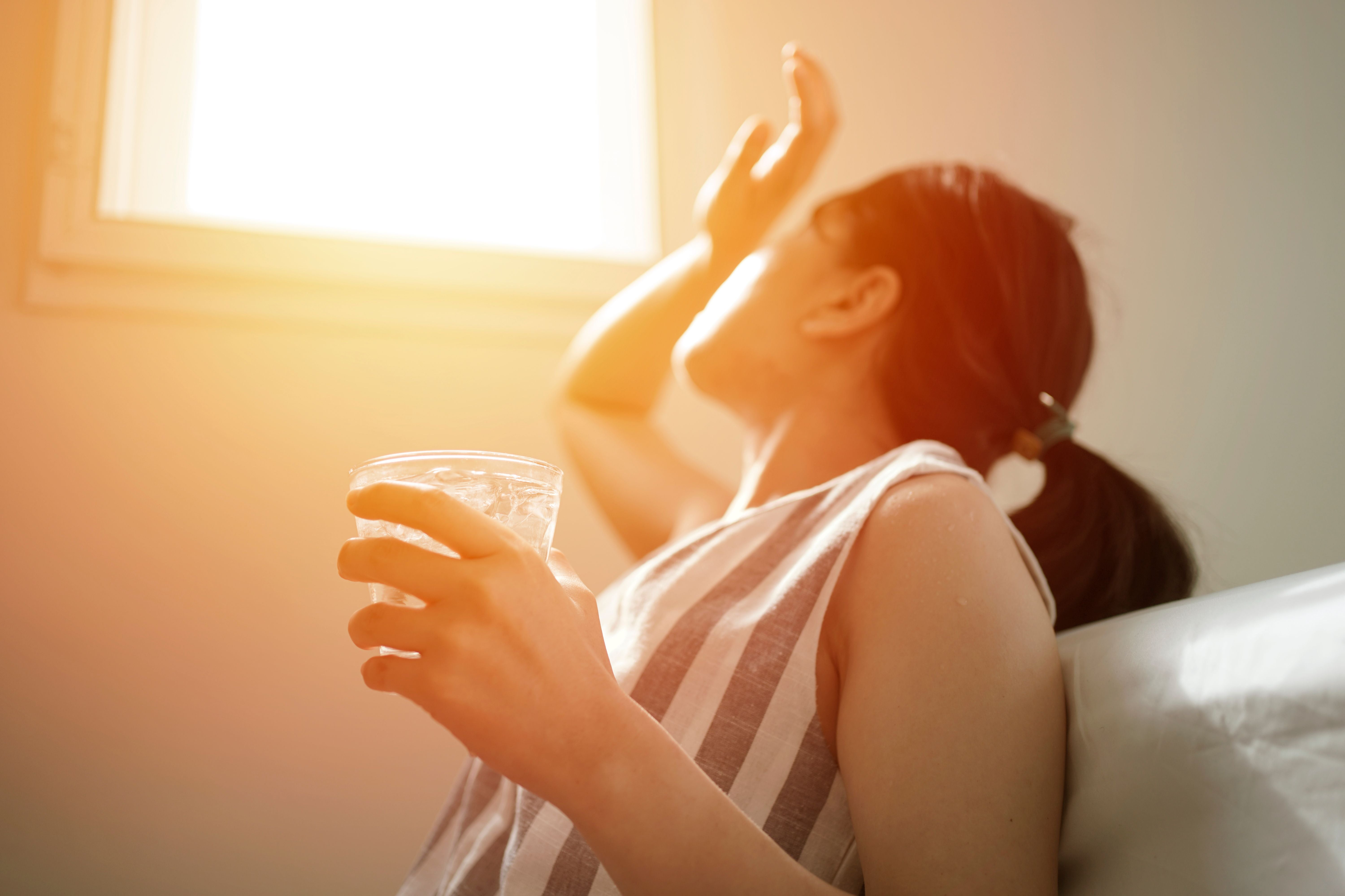 Frau schützt sich vor grellem Sonnenlicht am Fenster und hält ein Glas Wasser mit Eis.