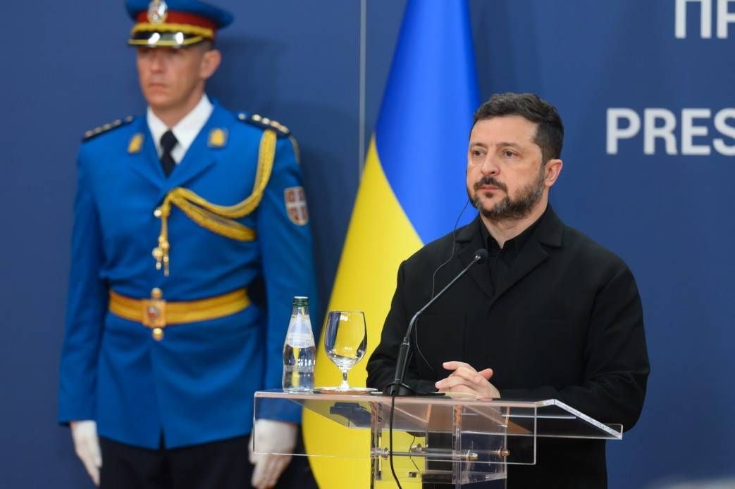 Ein Mann hält eine Rede am Rednerpult, hinter ihm steht ein uniformierter Wachmann und eine ukrainische Flagge.