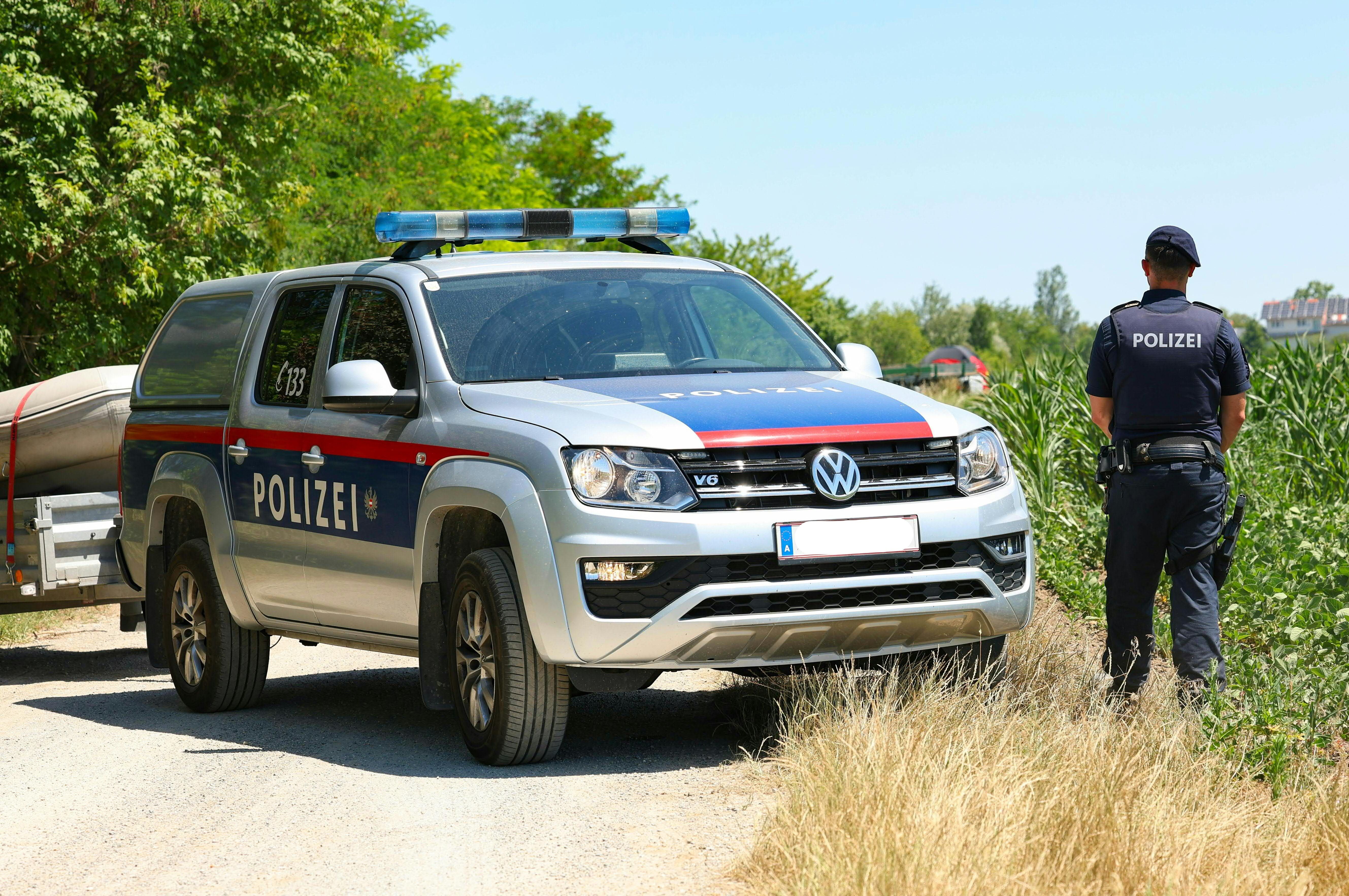 Ein österreichisches Polizeiauto und ein Polizist stehen an einem Feldweg neben dichtem Gras.