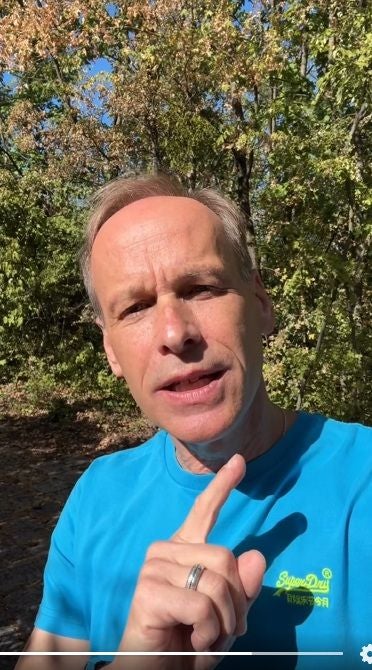 Ein Mann im blauen T-Shirt steht im Sonnenschein vor grünen Bäumen und spricht in die Kamera.