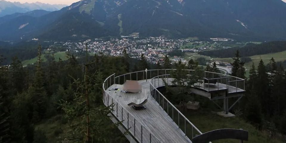 Holz-Aussichtsplattform im Wald mit Blick auf ein Tal und eine Stadt vor Bergkulisse.