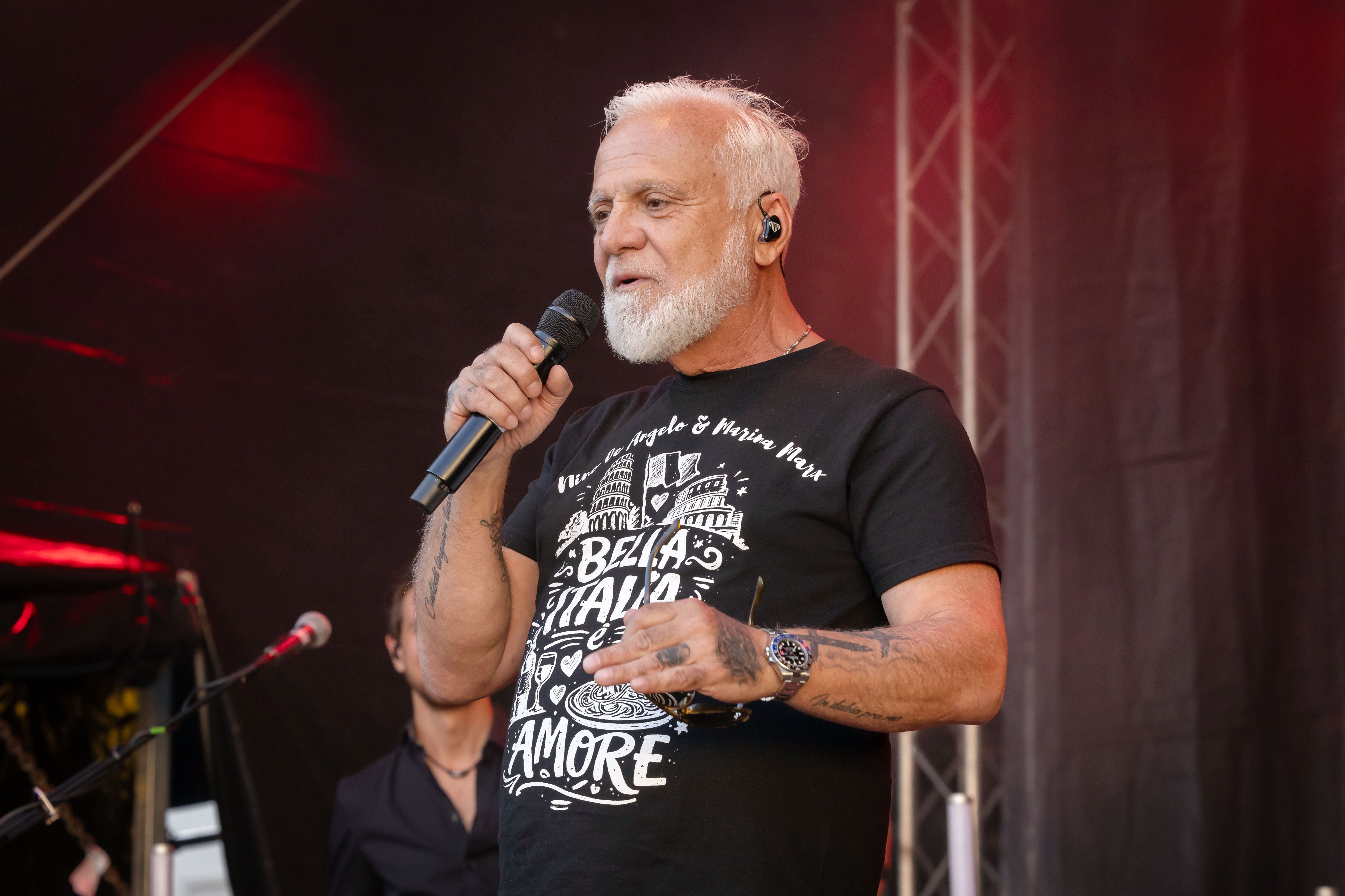 Ein älterer Mann mit Mikrofon singt auf einer Konzertbühne vor dunklem Hintergrund.
