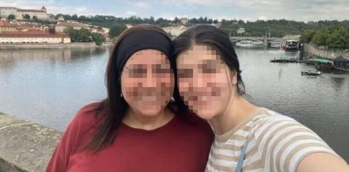 Zwei Frauen machen ein Selfie vor einem Fluss mit Brücke und Stadt im Hintergrund.