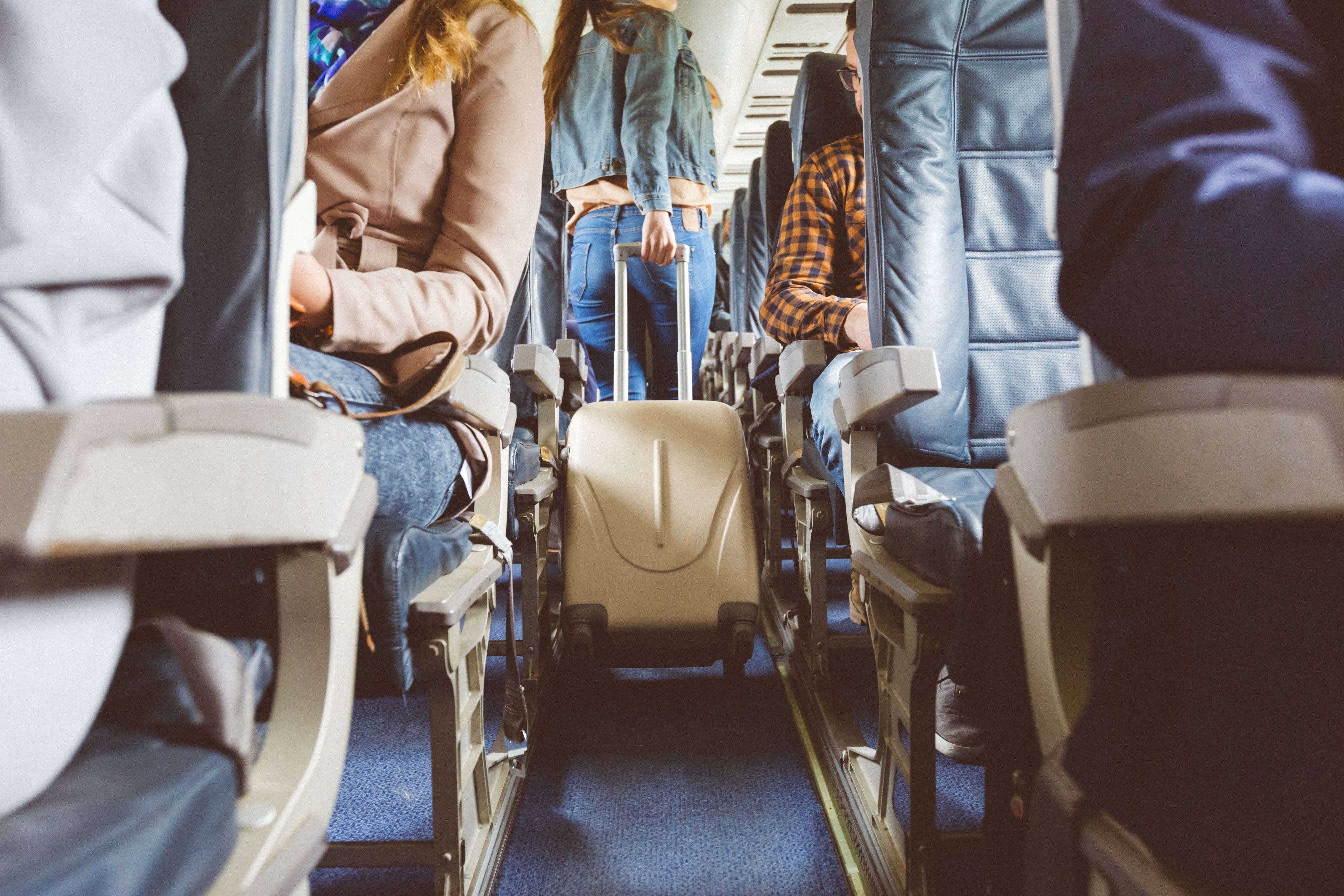 Passagier mit Koffer geht durch den Gang eines Flugzeugs, andere sitzen bereits auf ihren Plätzen.