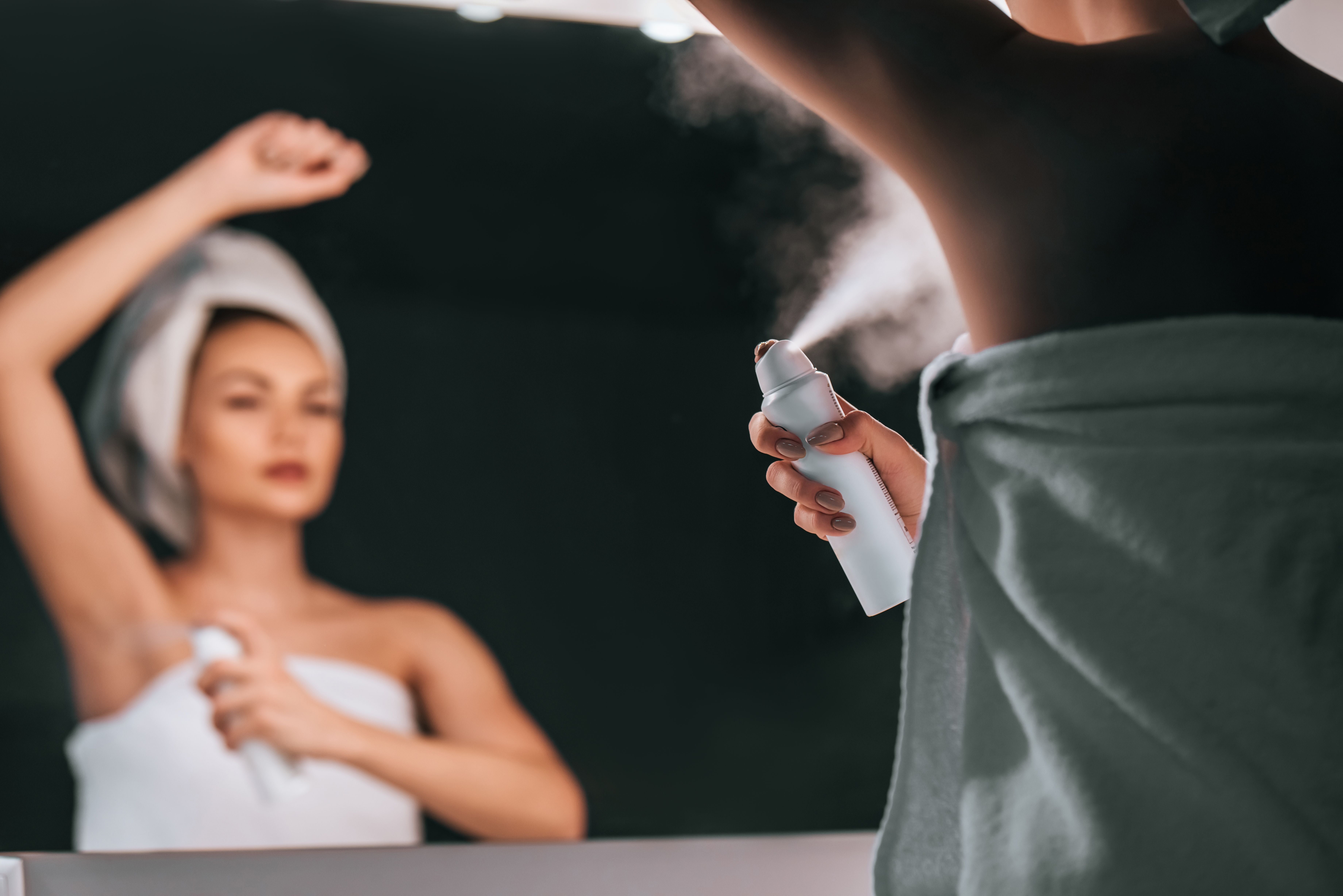 Frau im Handtuch sprüht Deodorant unter die Achseln vor einem Badezimmerspiegel.