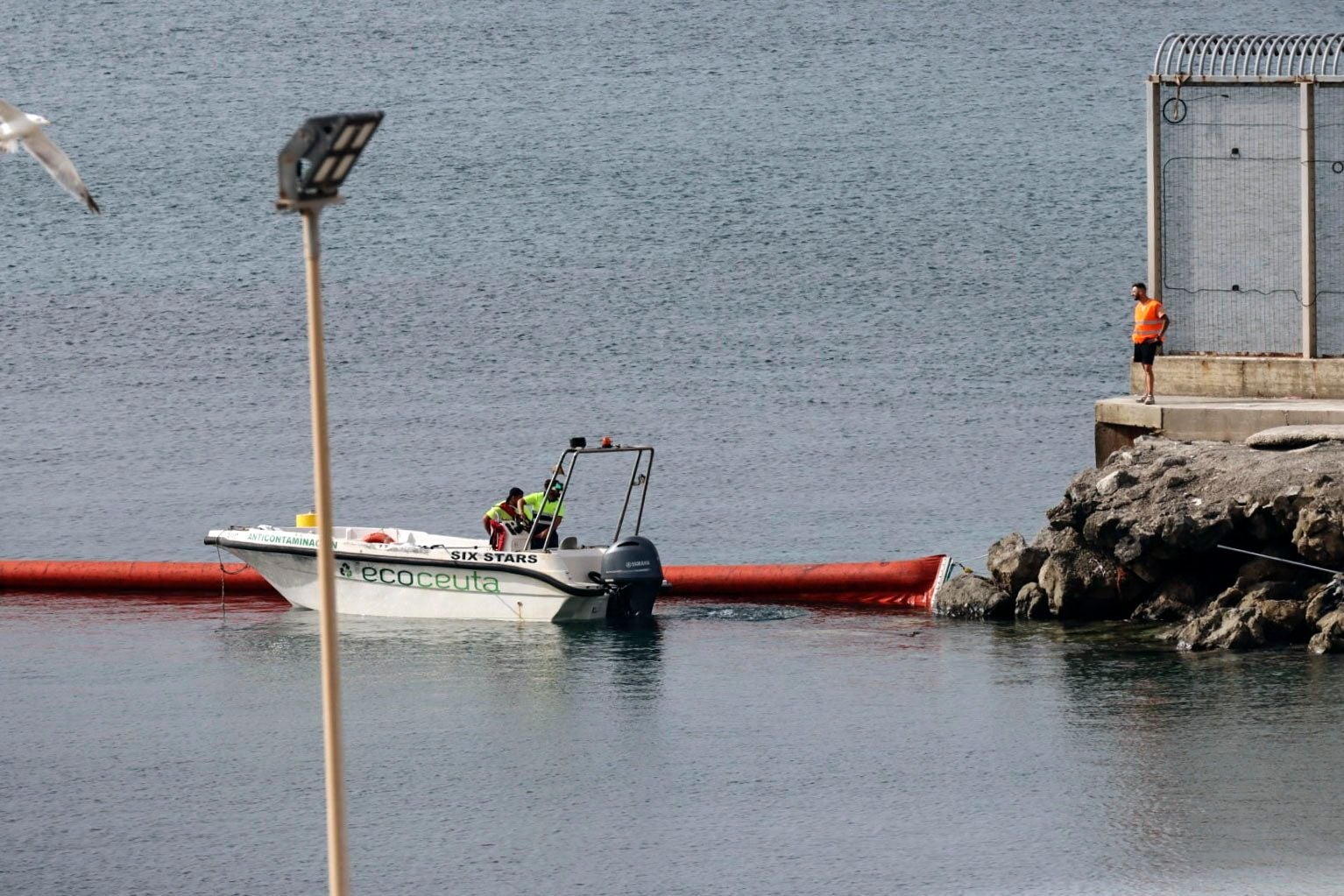 Ein Boot arbeitet an einer roten Schwimmbarriere im Meer bei Felsen und einem Zaun an der Küste.