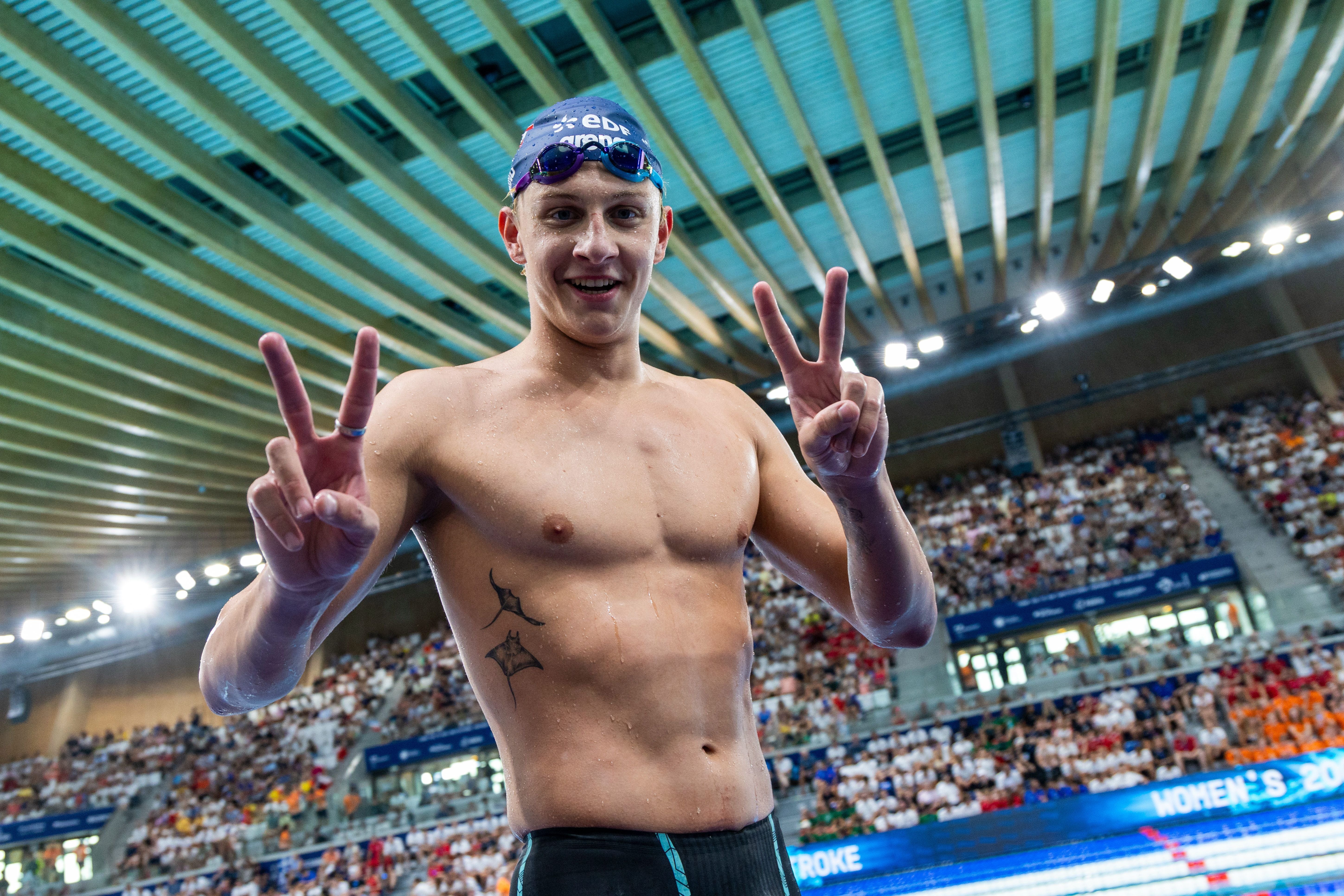 Ein Schwimmer posiert lächelnd mit Victory-Zeichen vor einer vollen Schwimmhalle.