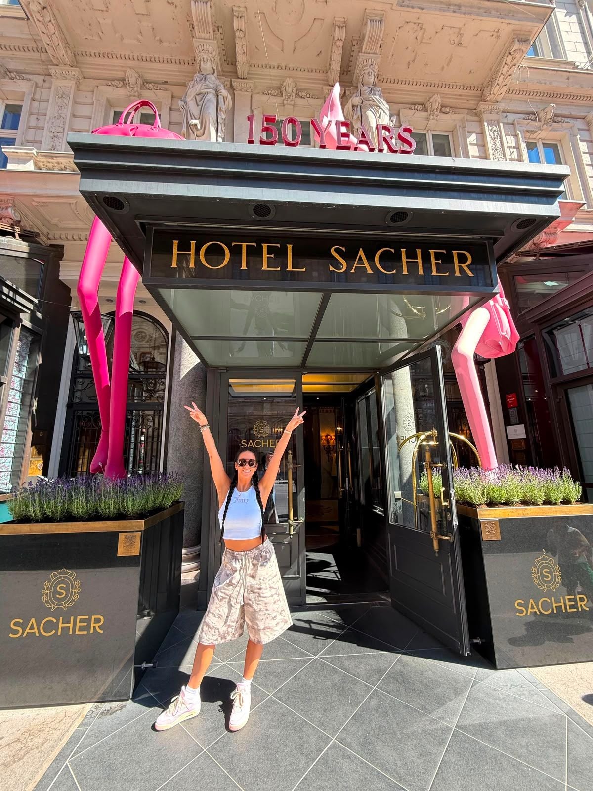 Frau posiert vor dem Eingang des Hotel Sacher, darüber steht „150 Years“ in roten Buchstaben.