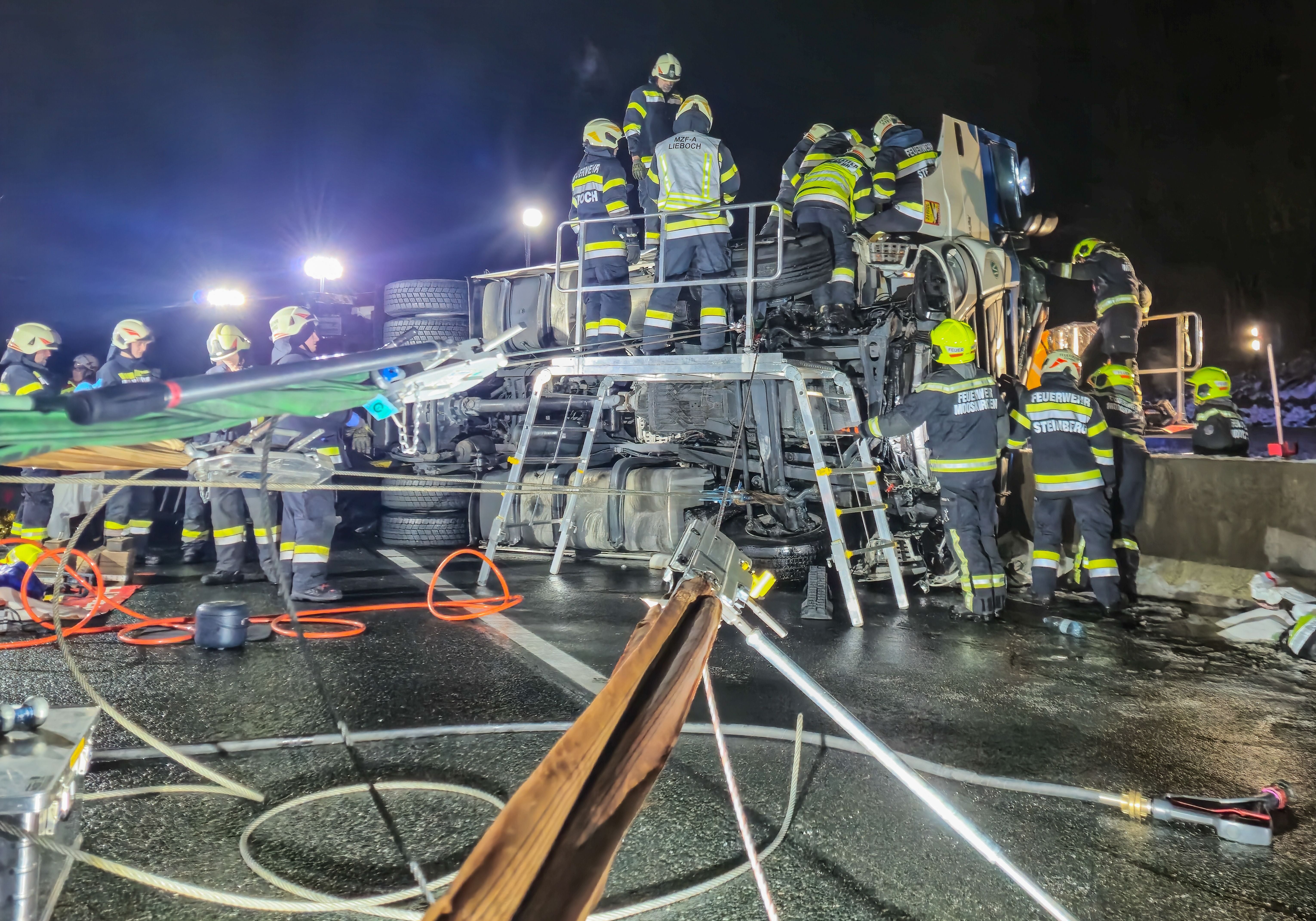 Feuerwehrleute bergen nachts einen umgestürzten LKW auf einer nassen Autobahn.